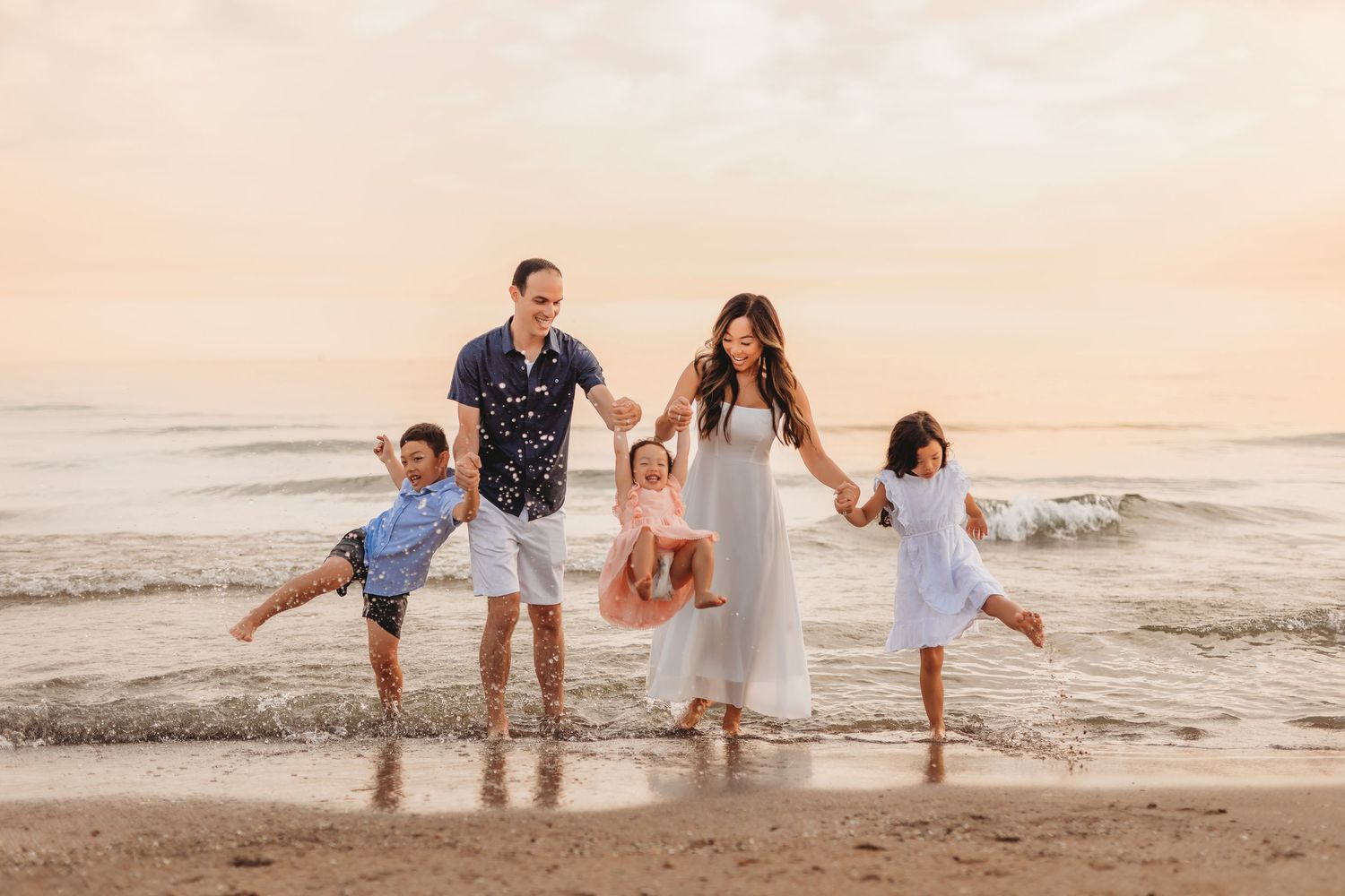 Family of five joyfully playing on a sandy beach at sunset, holding hands and swinging children near ocean waves.