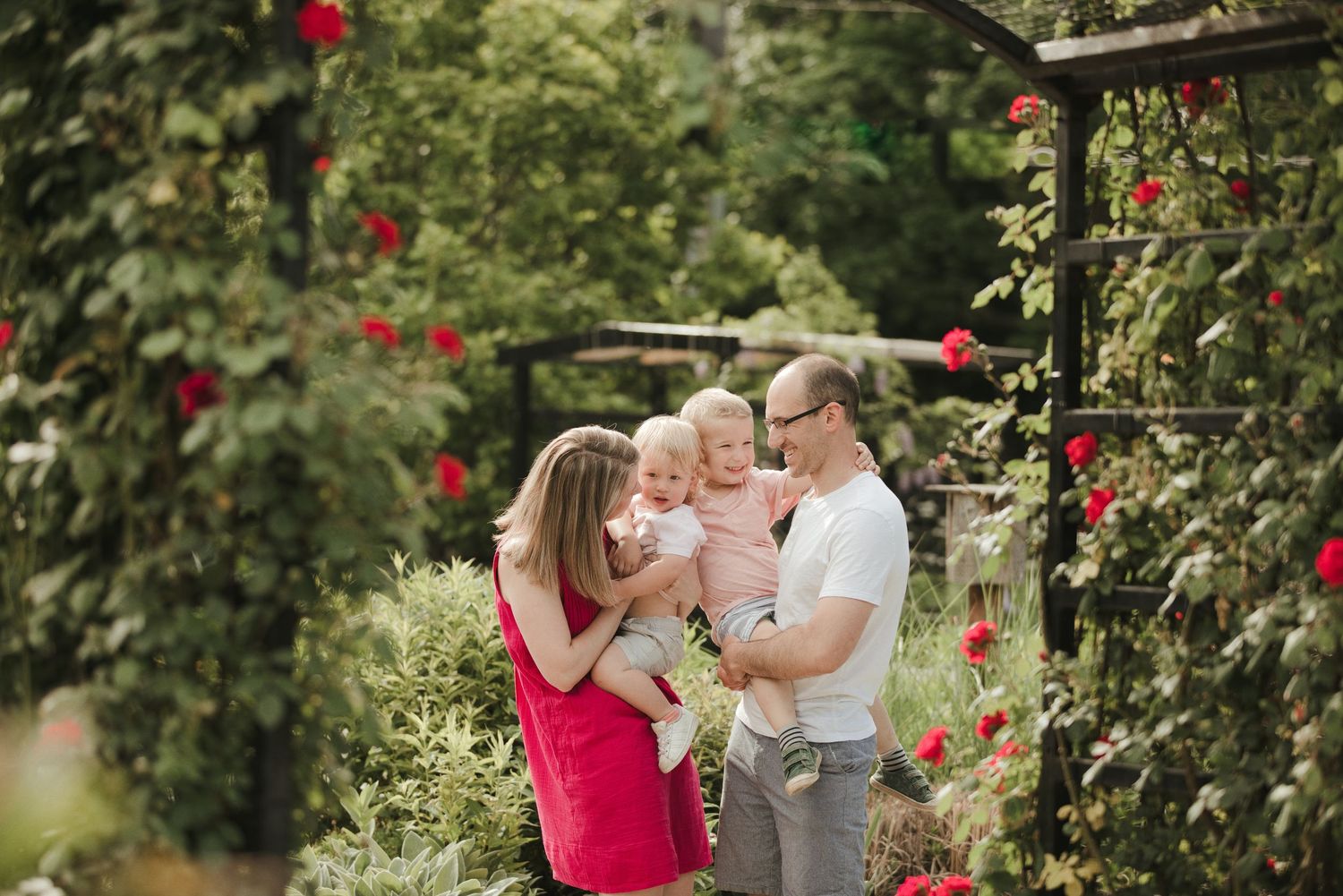 Family with baby enjoying a garden surrounded by red roses and lush greenery on a sunny day.