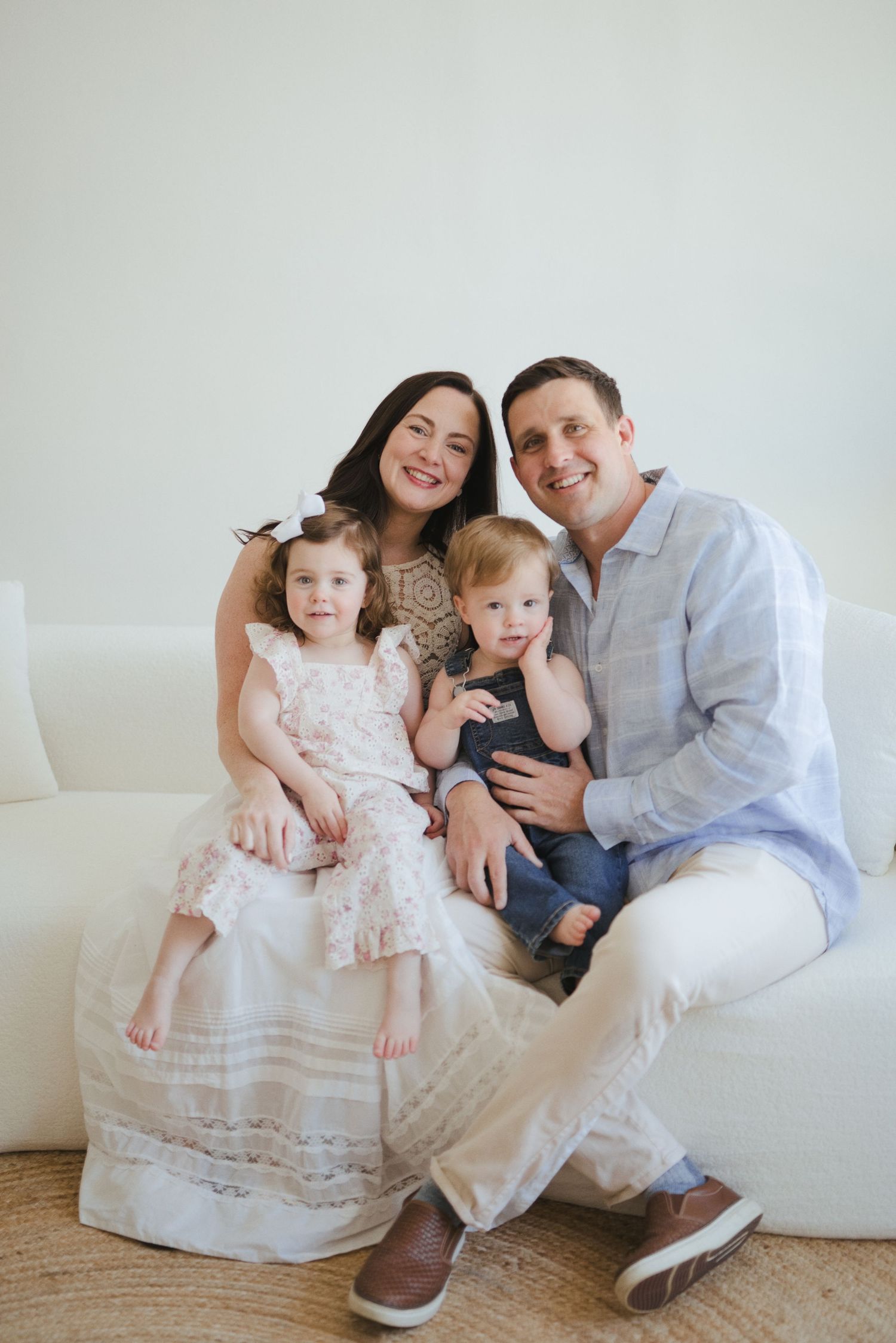 Happy family of four posing on white sofa, parents smiling with toddler girl in pink dress and baby boy in jeans.