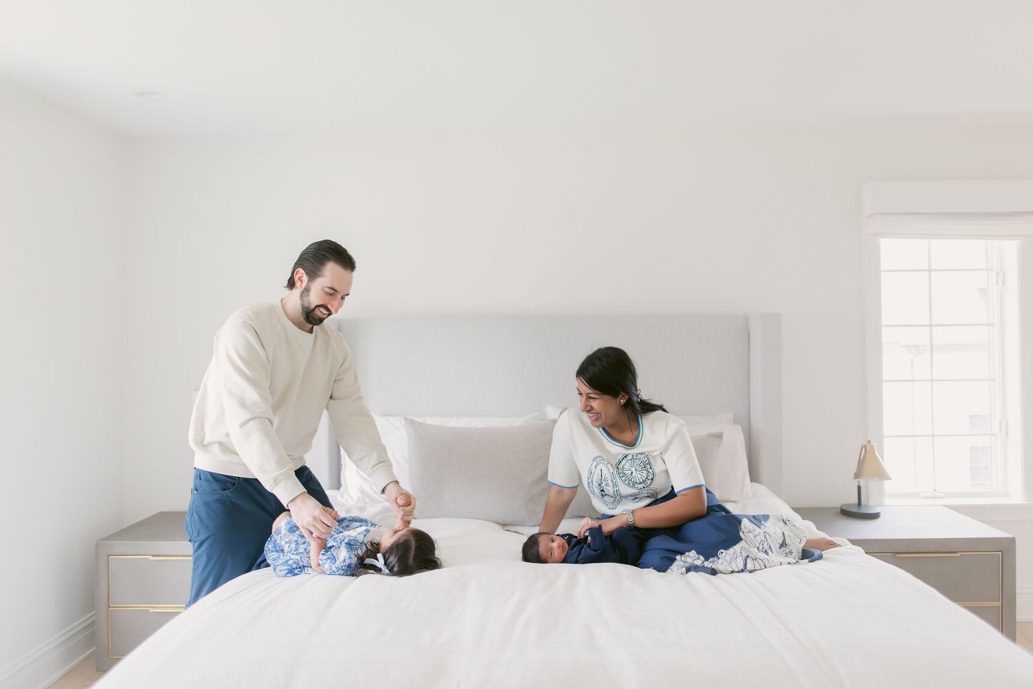 Family playing on a white bed in a bright minimalist bedroom, parents smiling at baby lying down.