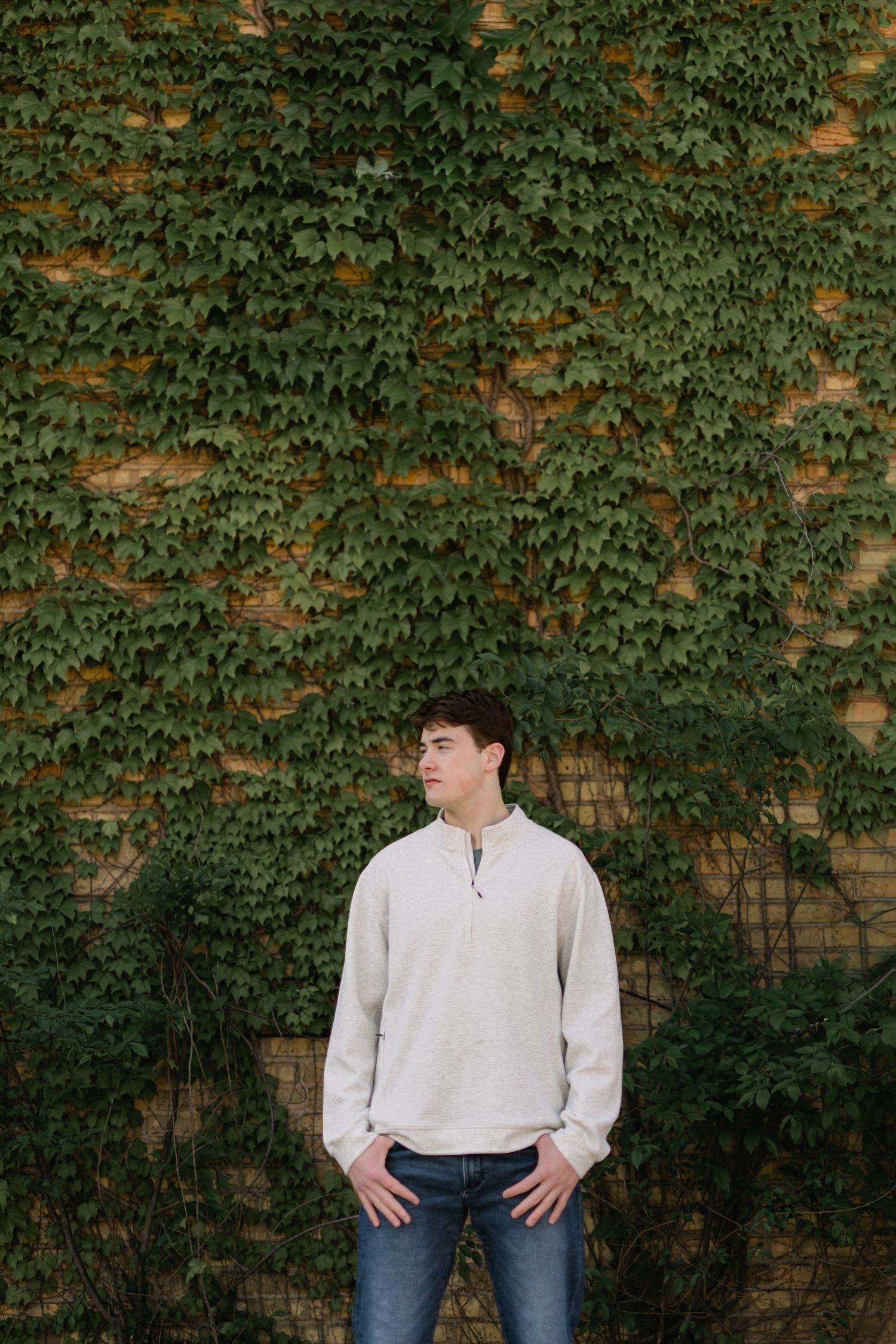 Young man in cream quarter-zip sweater and jeans standing against ivy-covered brick wall outdoors.
