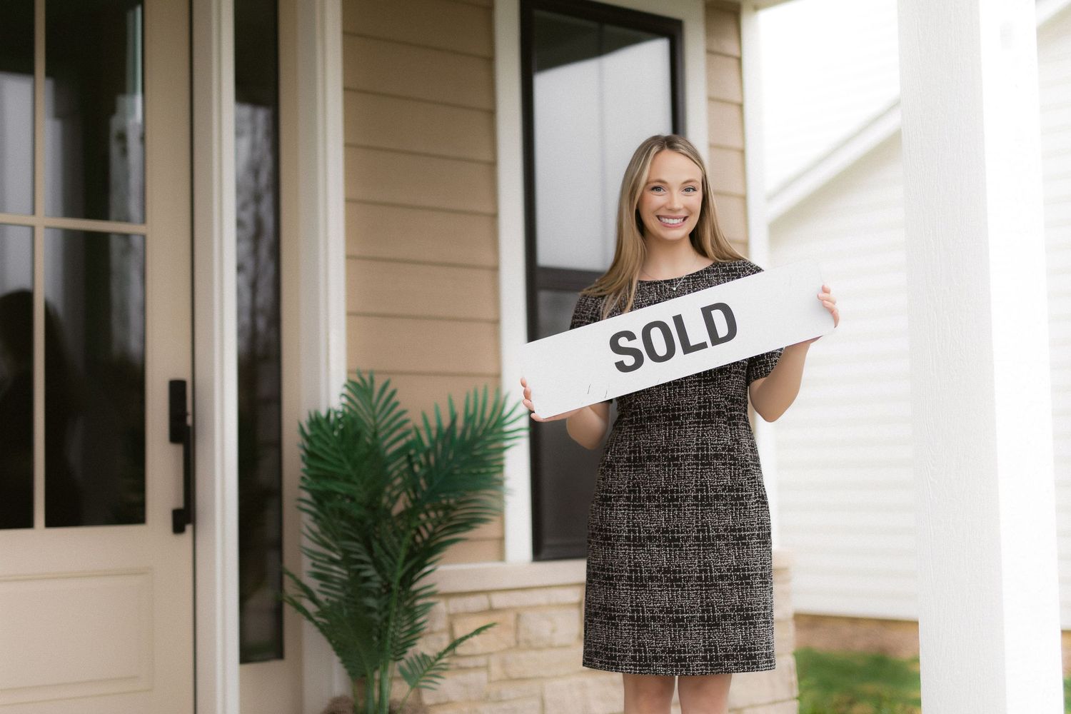 Real estate professional displays 'Sold' sign while standing on front porch with palm plant.