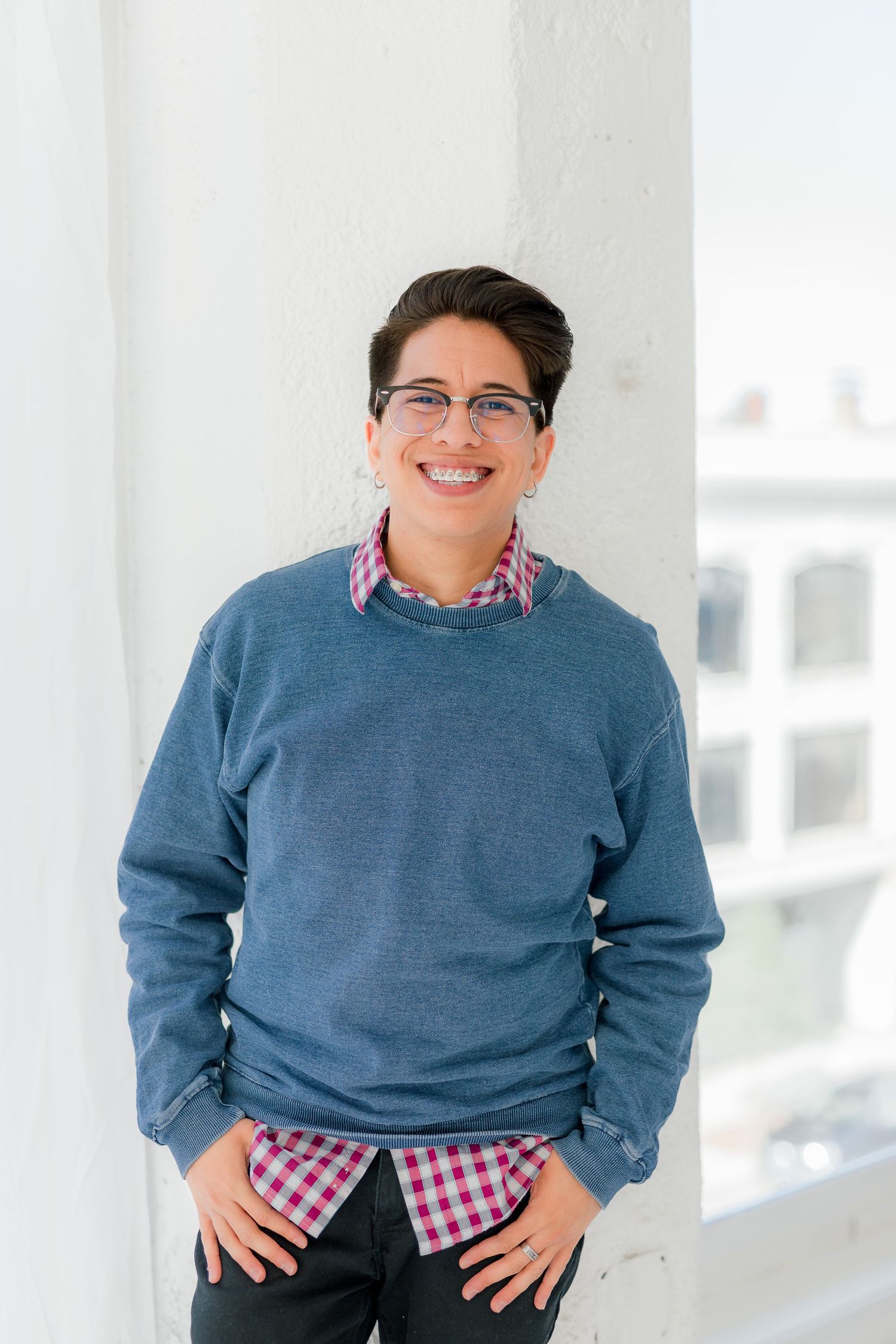 Portrait of someone in blue sweater and checkered shirt smiling against white column background.