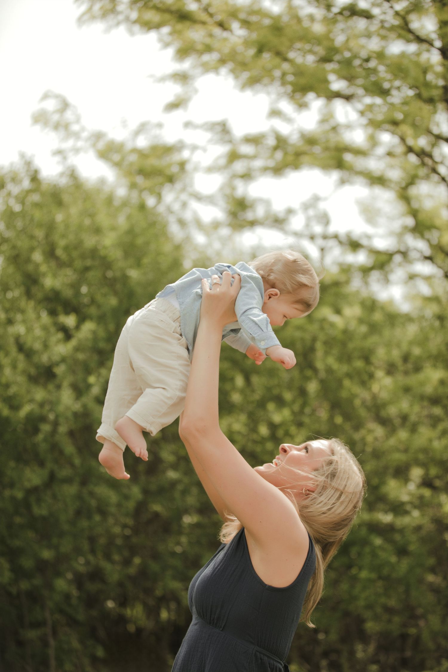 A joyful mother lifts her smiling baby high in the air outdoors, surrounded by lush green trees on a sunny day.