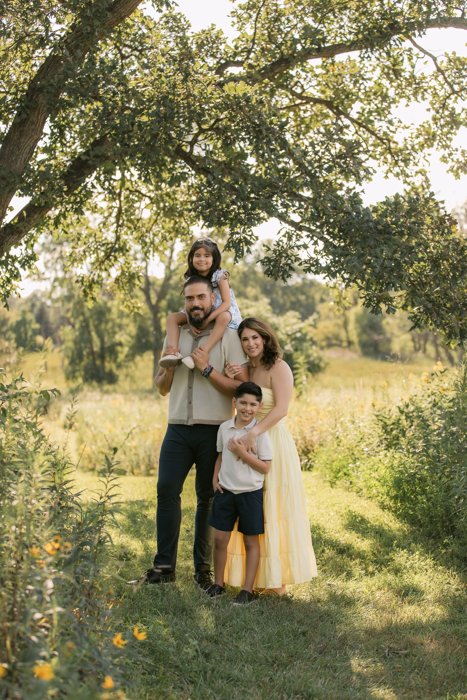 A family shares joyful moments together in a sun-dappled meadow under tall trees during a golden hour photoshoot.