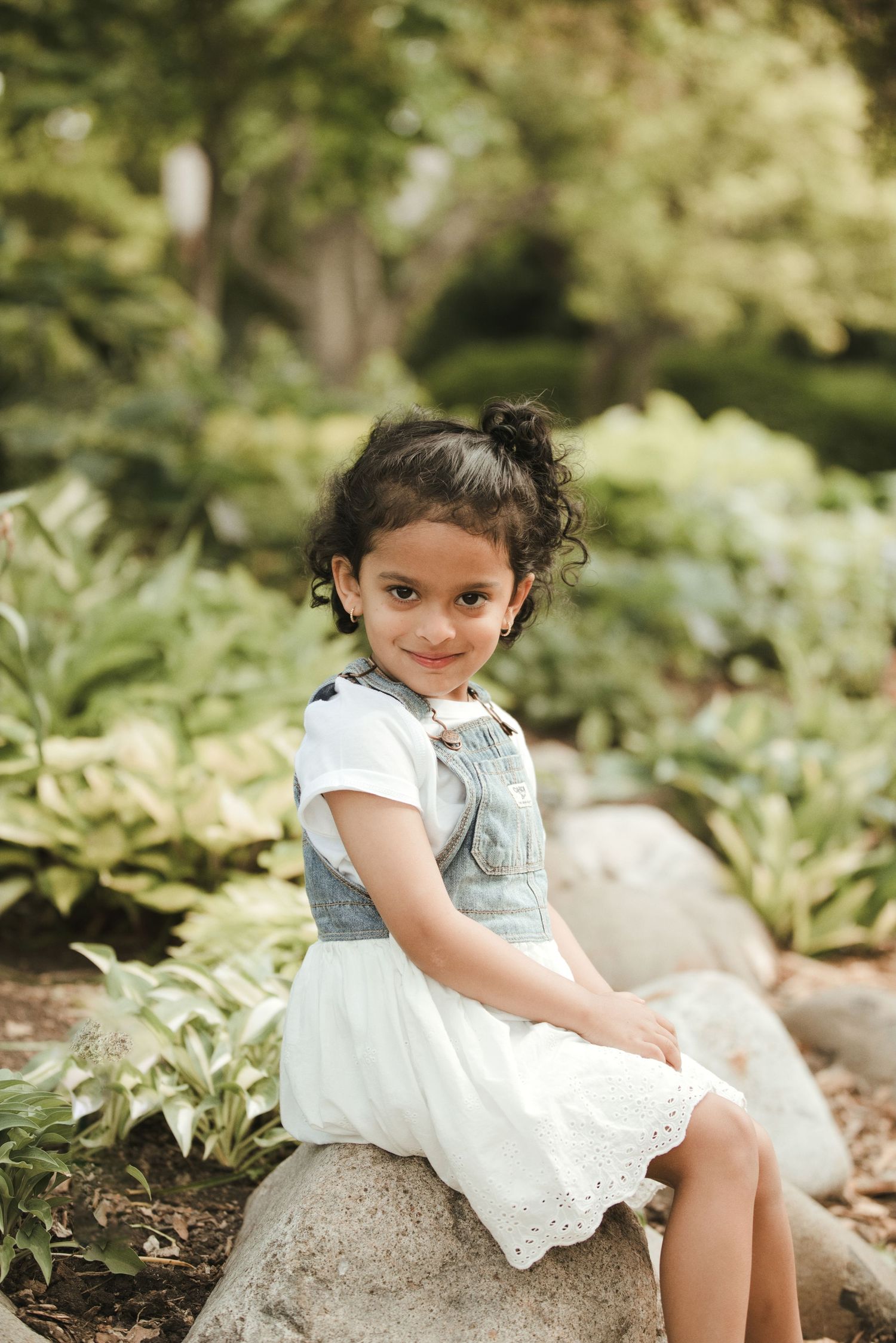 Young girl in white dress and denim vest sitting on rocks in a lush garden, smiling at camera.