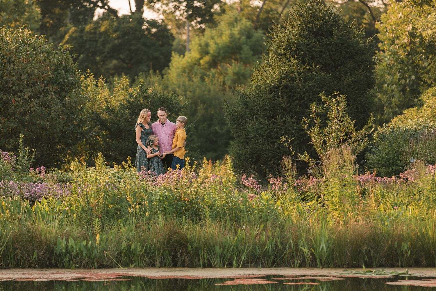 Family of three standing among wildflowers and lush greenery near a pond in golden hour sunlight.