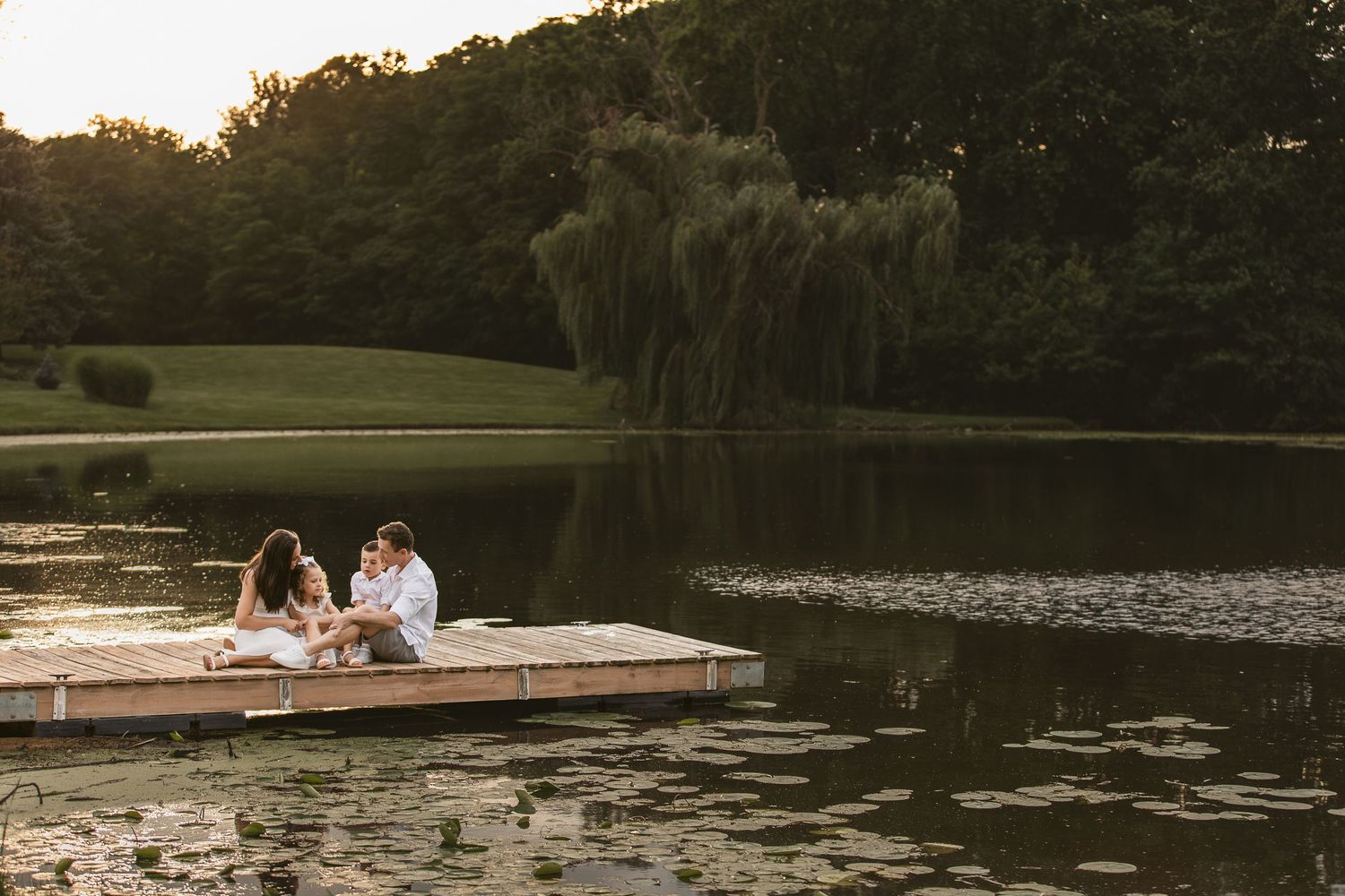Couple sitting on a wooden dock by a serene lake at sunset, surrounded by lush green trees and weeping willows.