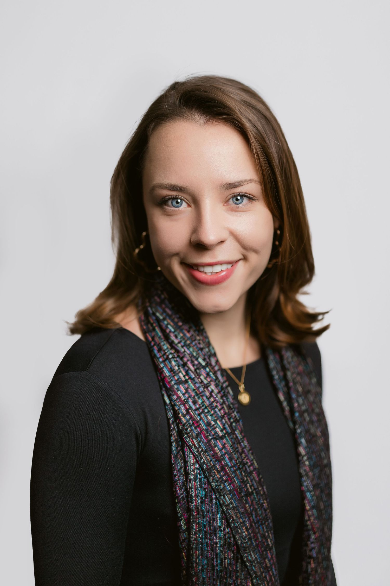 Professional headshot of person wearing black blazer and patterned scarf against grey background.