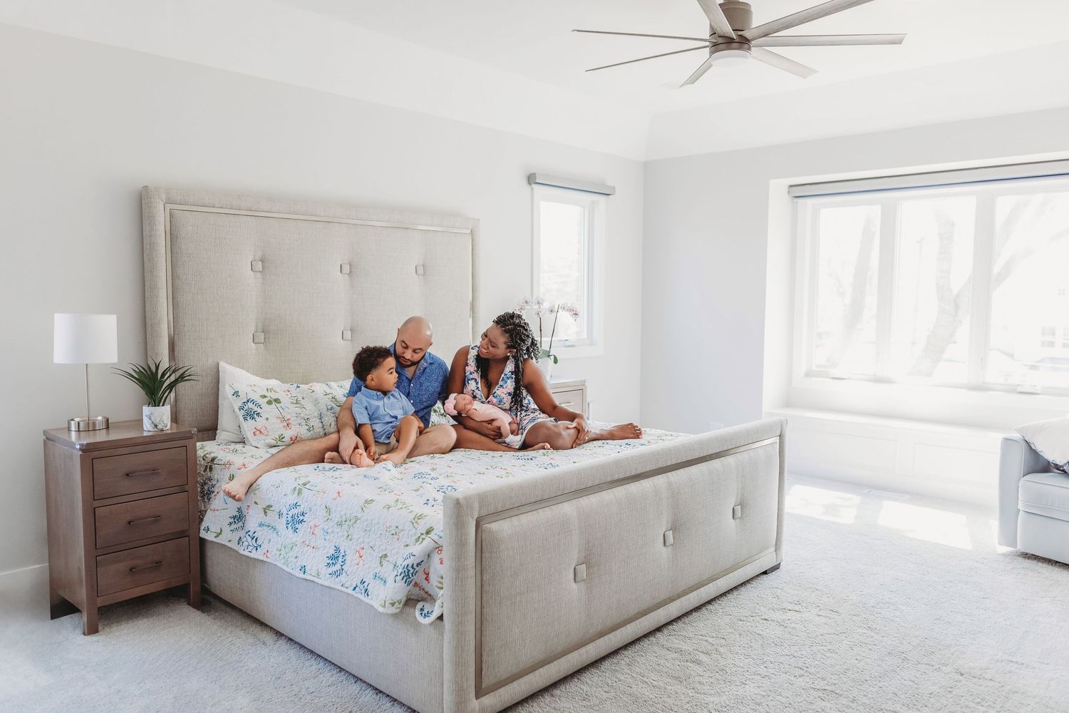 A light and airy bedroom features a tufted gray bed with floral bedding, wood nightstand, and a ceiling fan.