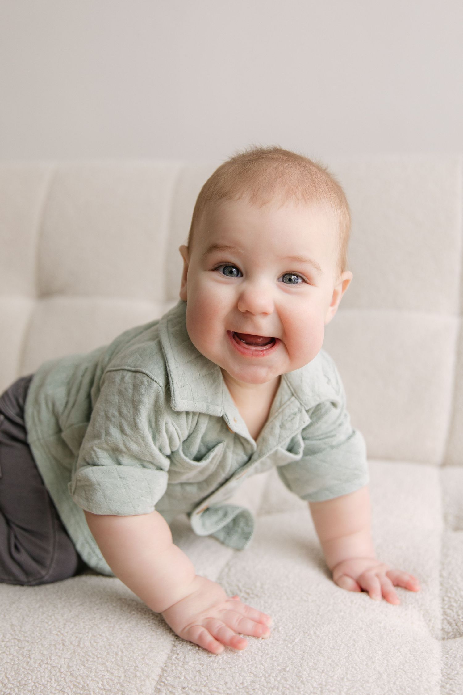 A happy baby in a mint green outfit lies on a white carpet while crawling and smiling.