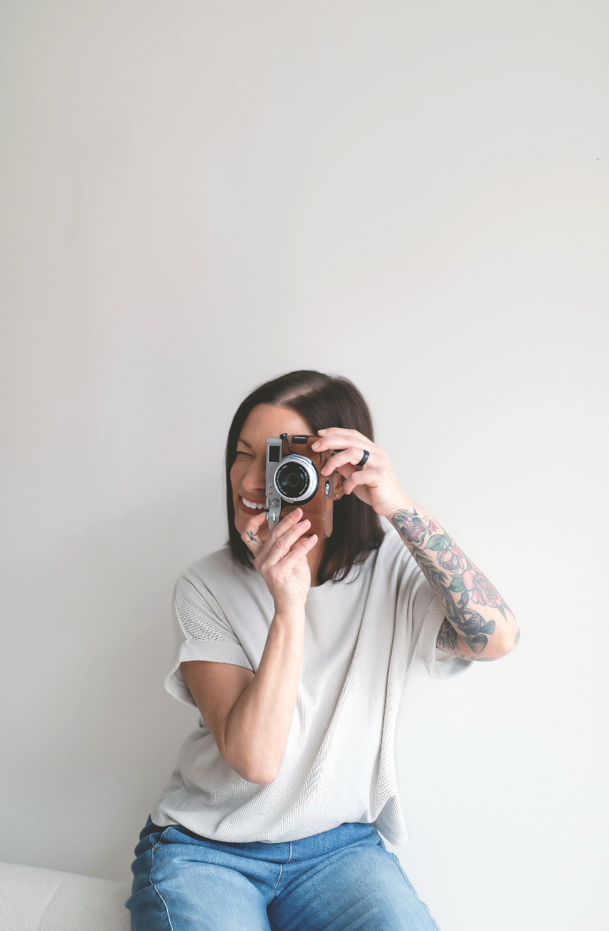 A smiling woman with a tattooed arm holds a vintage film camera up to her face, wearing a gray tee and jeans.