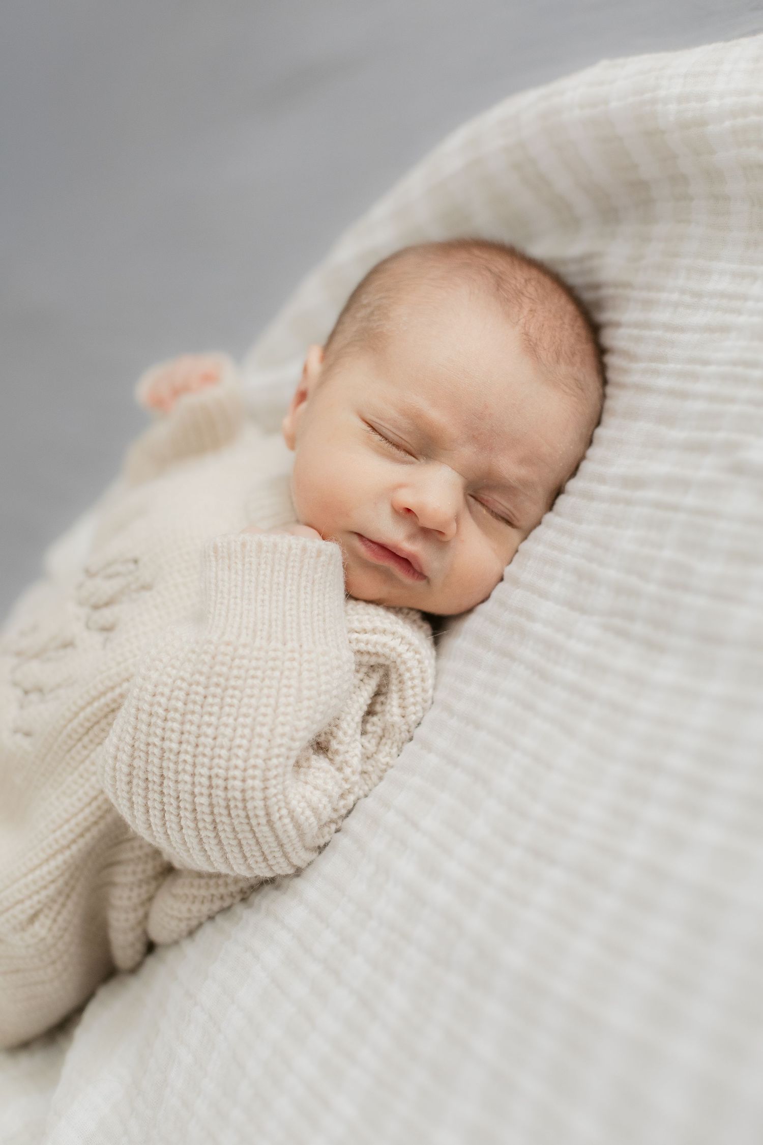 Sleeping newborn in soft white knit sweater on textured blanket.