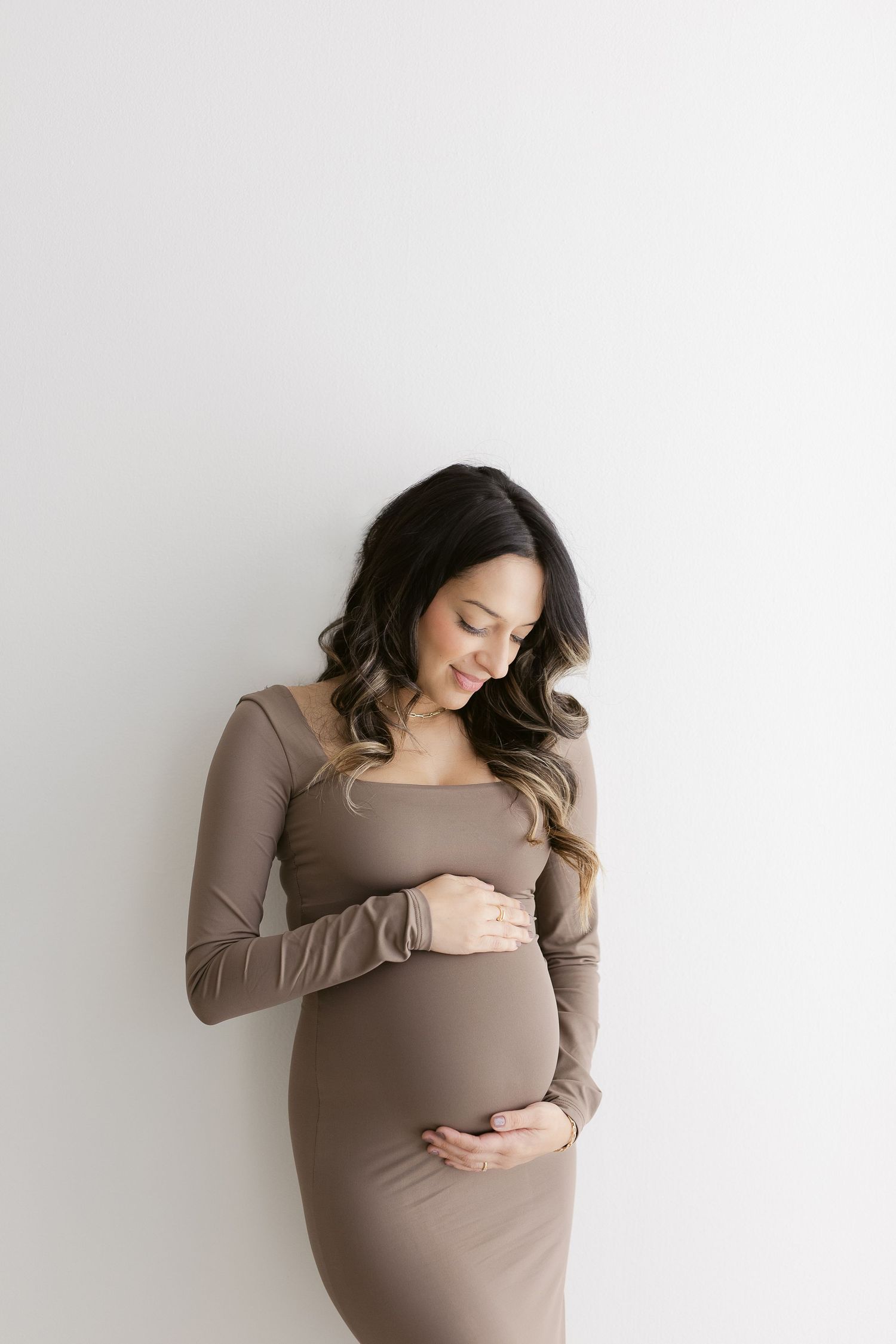 A pregnant woman in a brown fitted dress stands against a white wall, looking down with a gentle smile.