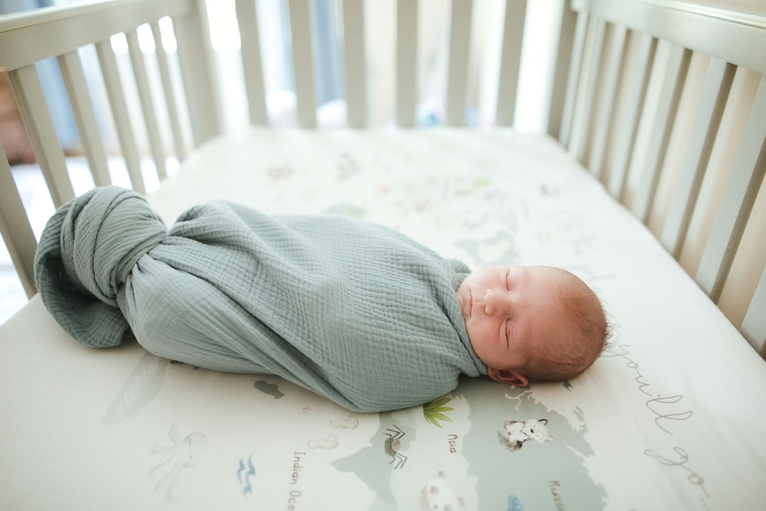 Newborn baby swaddled in sage green wrap sleeping peacefully in a white crib with a world map sheet.
