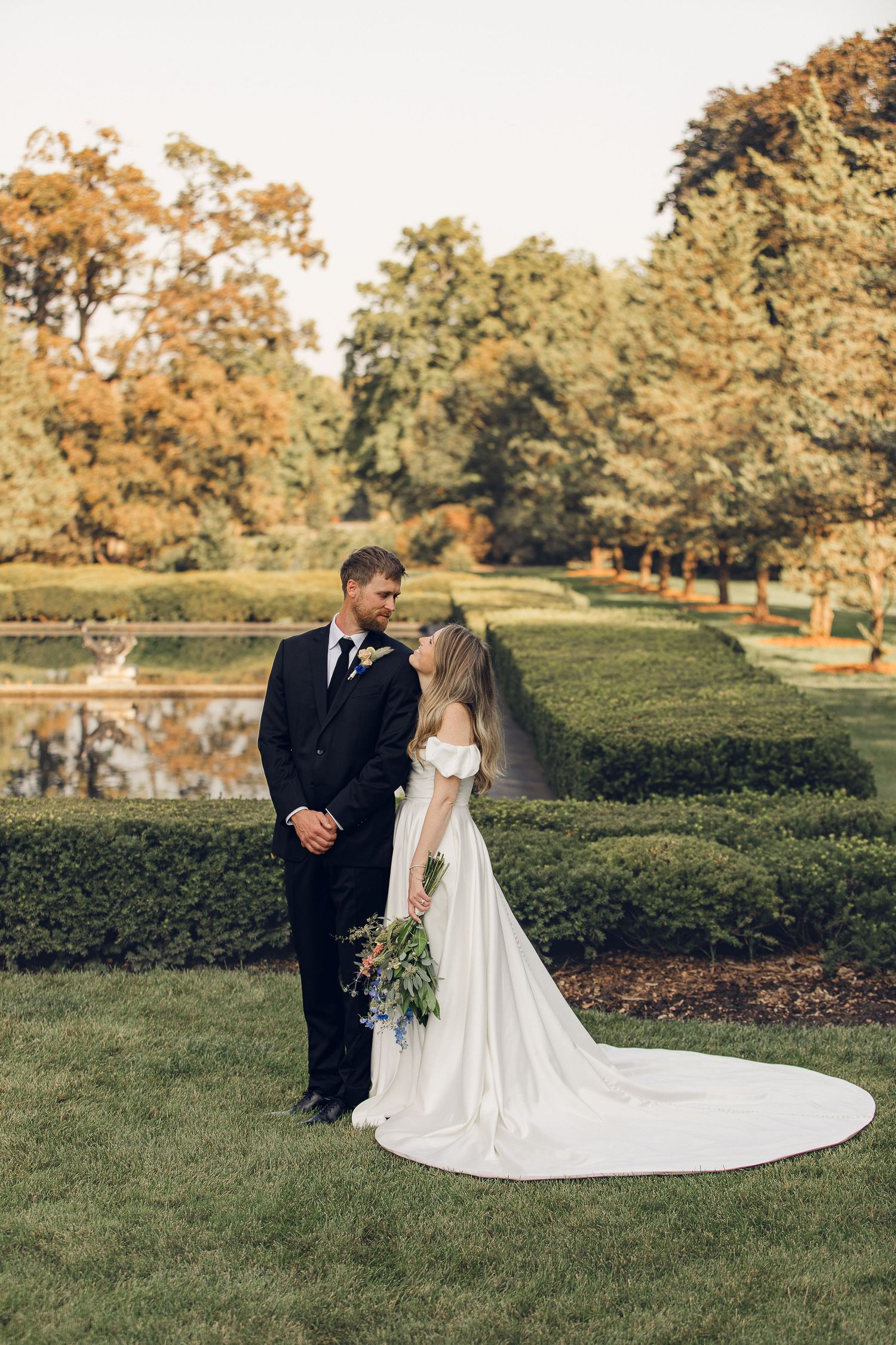 A romantic wedding couple stands together on manicured green lawn surrounded by autumn trees during their outdoor ceremony.
