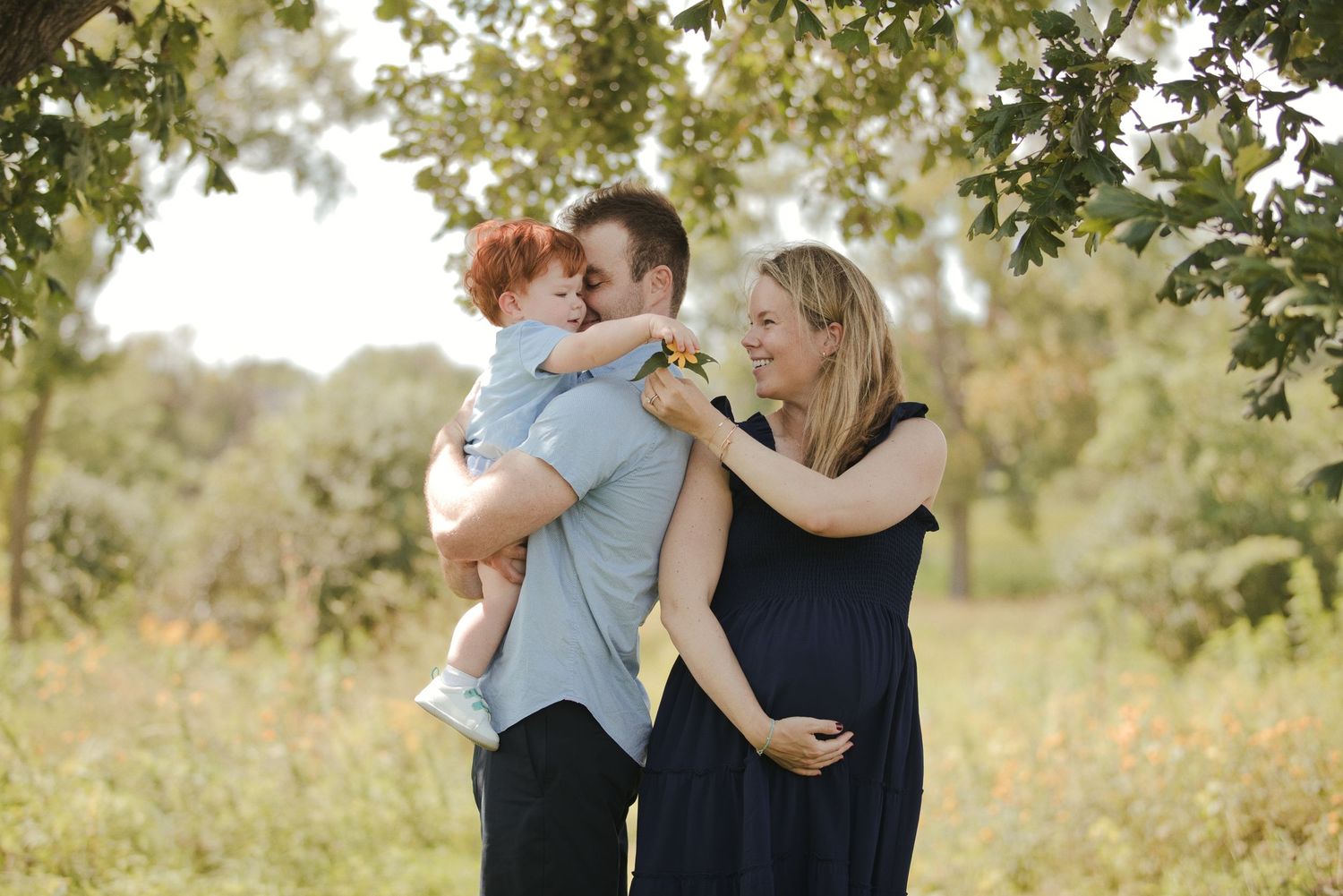 A happy family with a redheaded toddler and pregnant mother poses outdoors under green trees in warm sunlight.