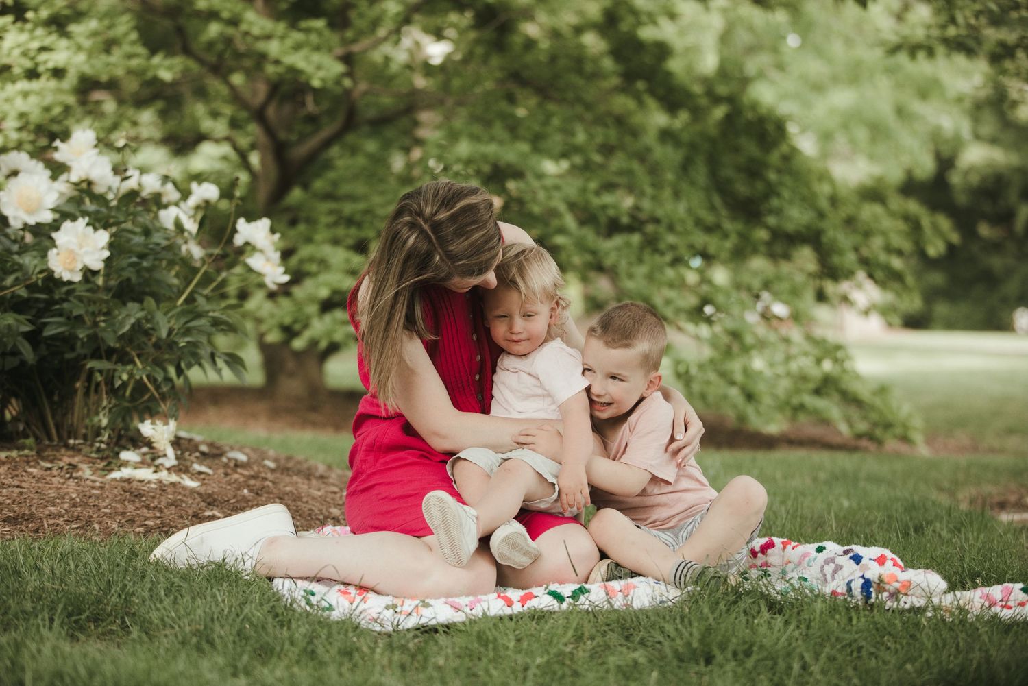 A mother in a red dress sits on a floral blanket in a garden, hugging two young children near blooming white flowers.