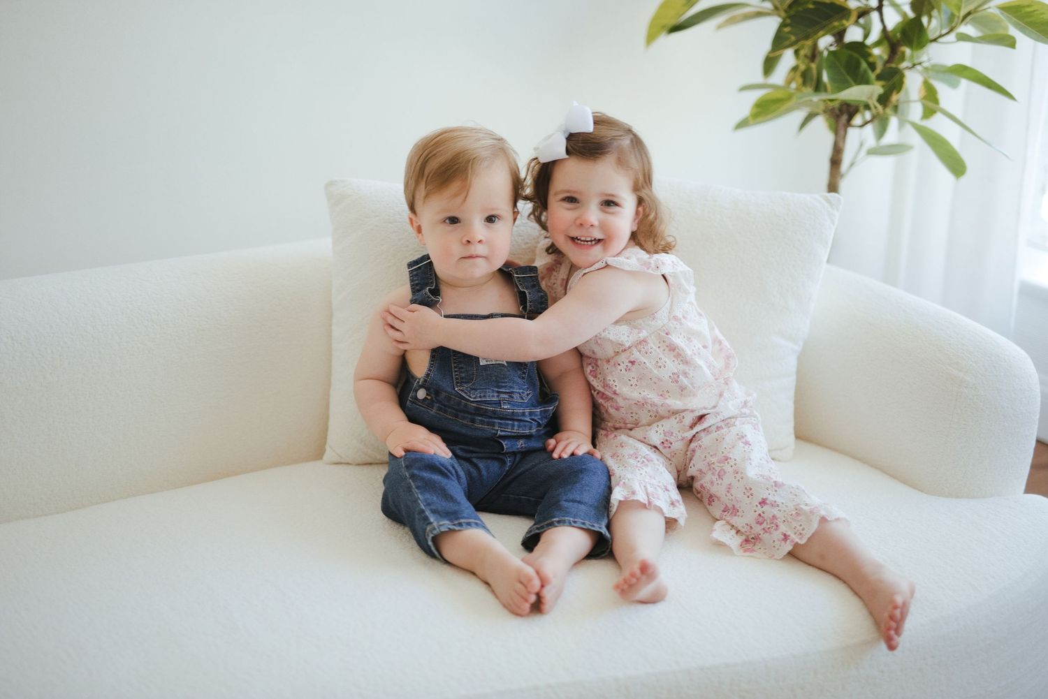 Two young siblings sitting on a white sofa, girl in floral dress hugging toddler boy in denim overalls.