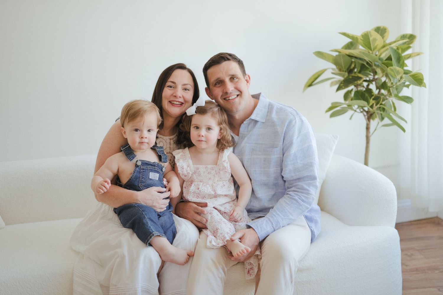 Happy family of four posing on white sofa, parents holding toddler boy and young girl in bright airy room.