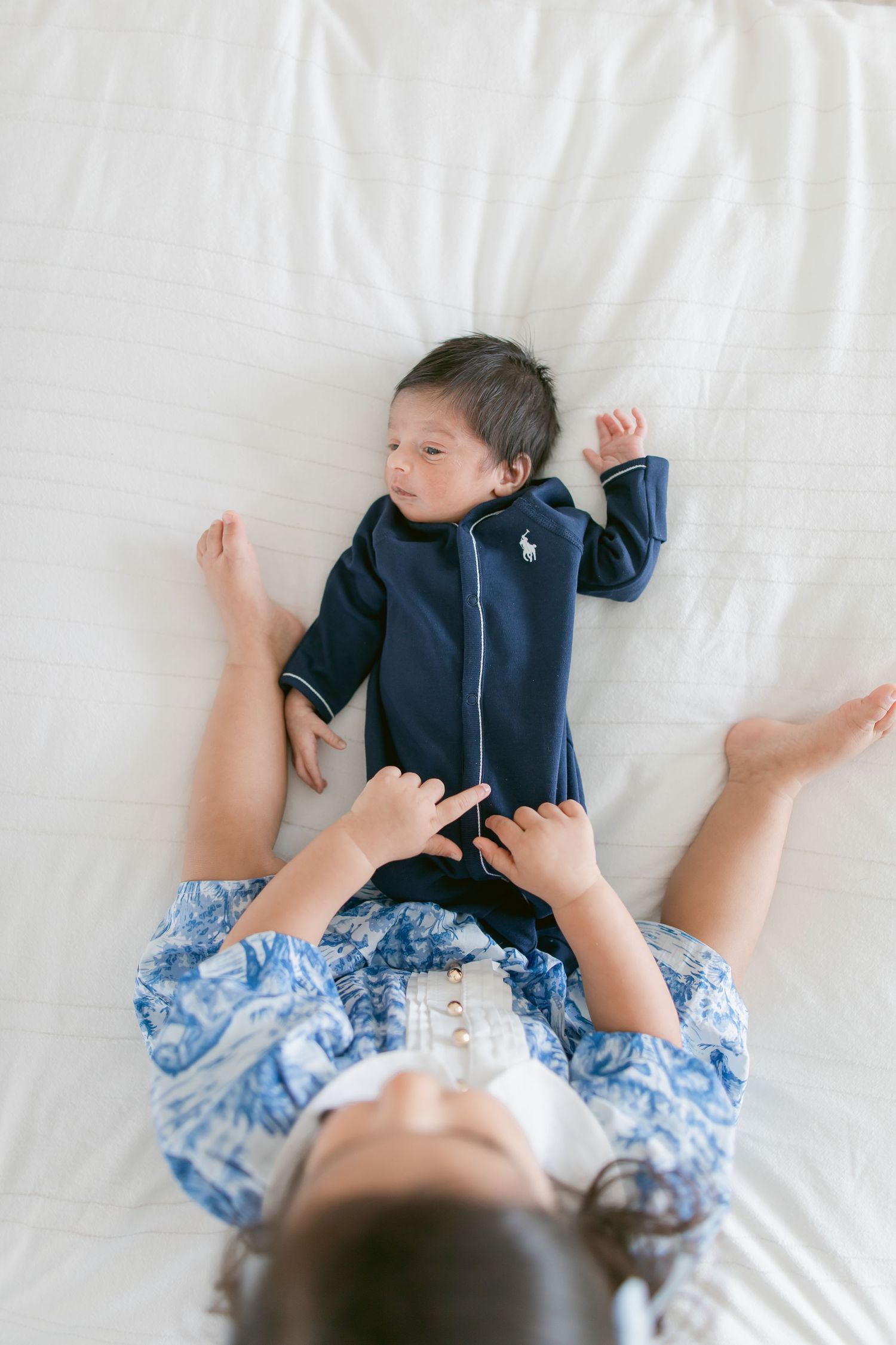 Overhead view of a toddler in blue floral outfit interacting with a newborn baby in navy Ralph Lauren sleeper on white bed.