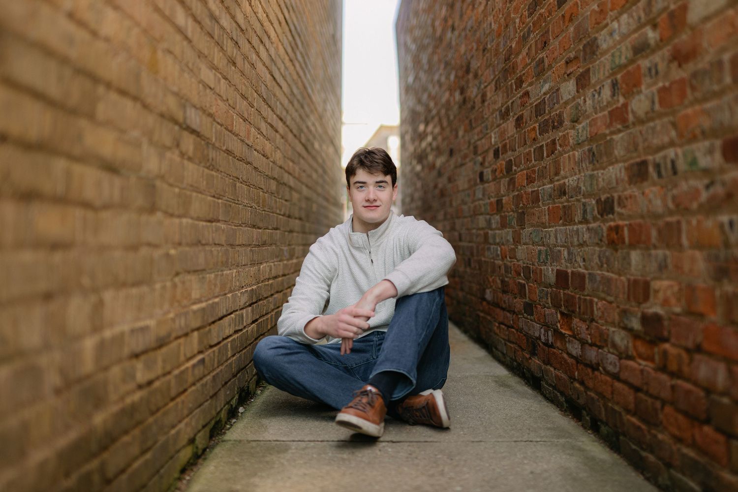 Young man sitting cross-legged in a narrow brick alleyway, wearing a light gray hoodie and blue jeans.