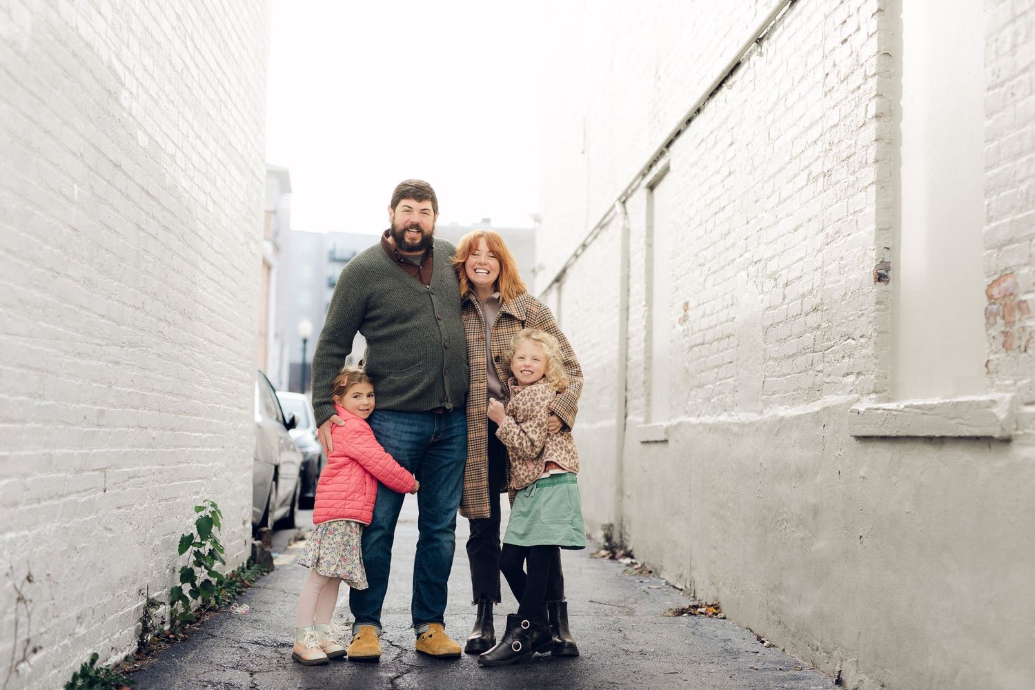A family in winter coats stands together in a white-walled alleyway on an overcast day.