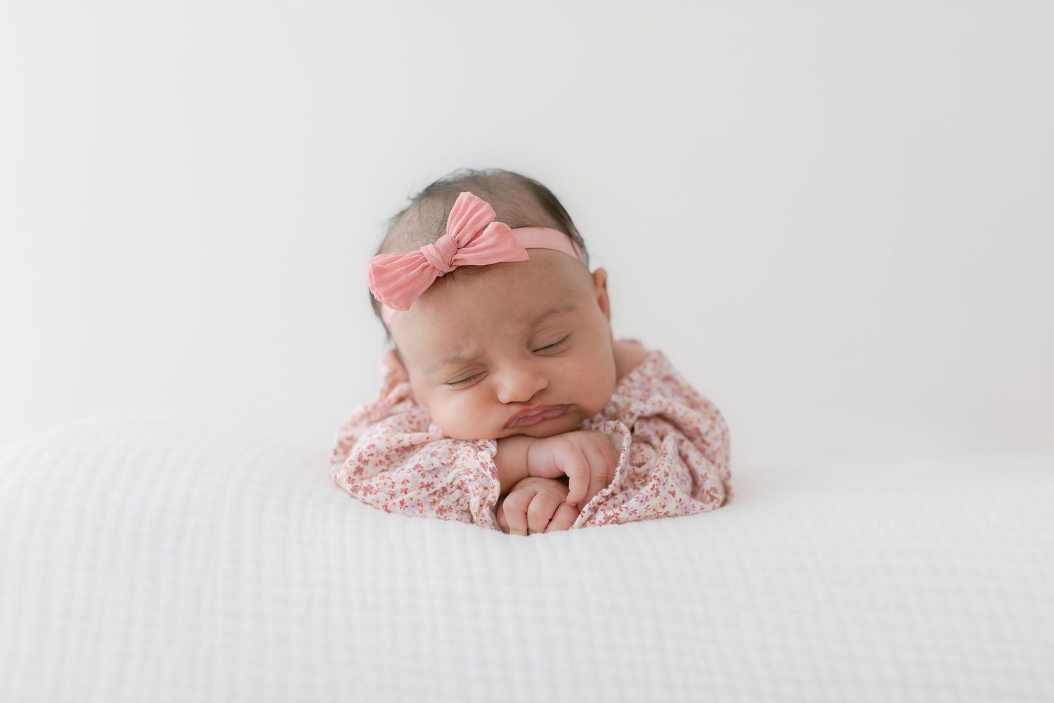 Sleeping newborn in pink outfit with bow headband on white background.