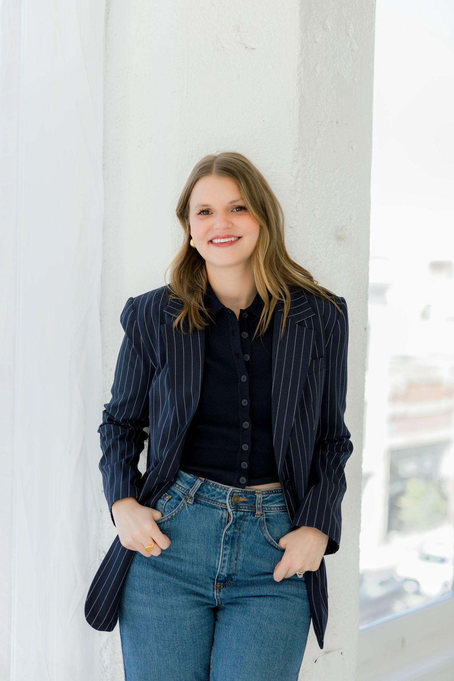 Professional portrait of someone in pinstriped blazer and jeans leaning against white column.