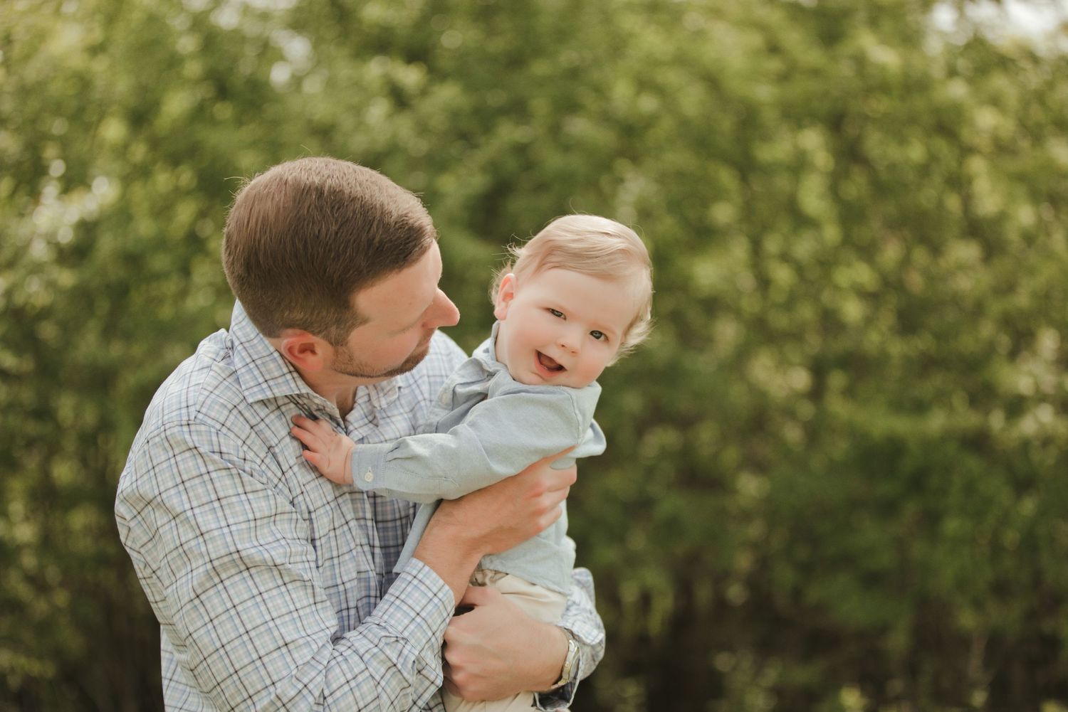 A father lovingly holds his smiling toddler outdoors with lush green trees in the background.