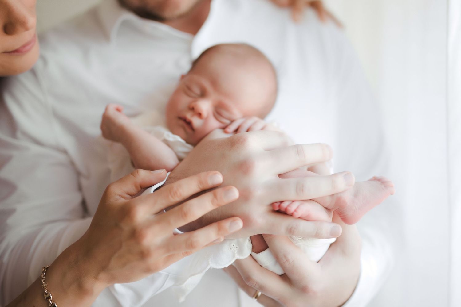Parents cradling a sleeping newborn baby in their hands, wearing white clothing in a soft, bright setting.