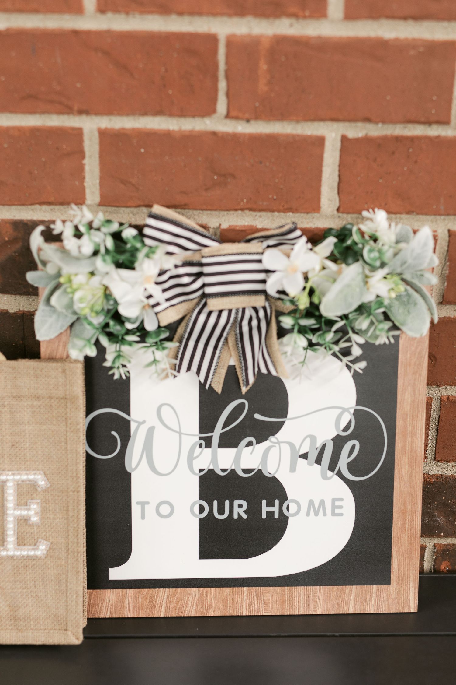 Decorative welcome sign with letter B, white flowers, and striped bow displayed against a brick wall.