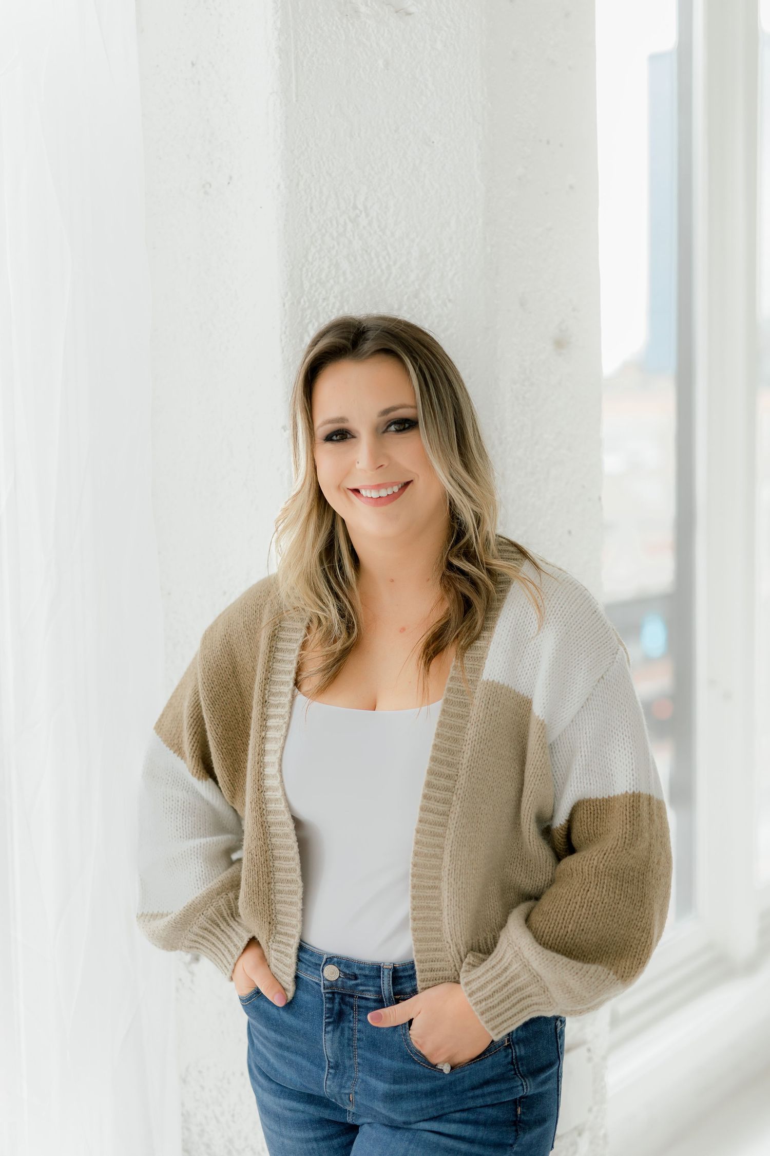 Model in beige cardigan and jeans poses against bright white studio background.