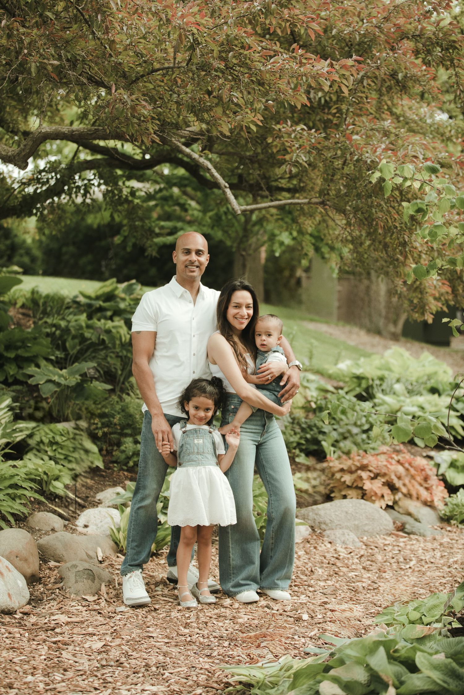 Family of four posing in a lush garden, parents with young daughter and baby, surrounded by green foliage and rocks.