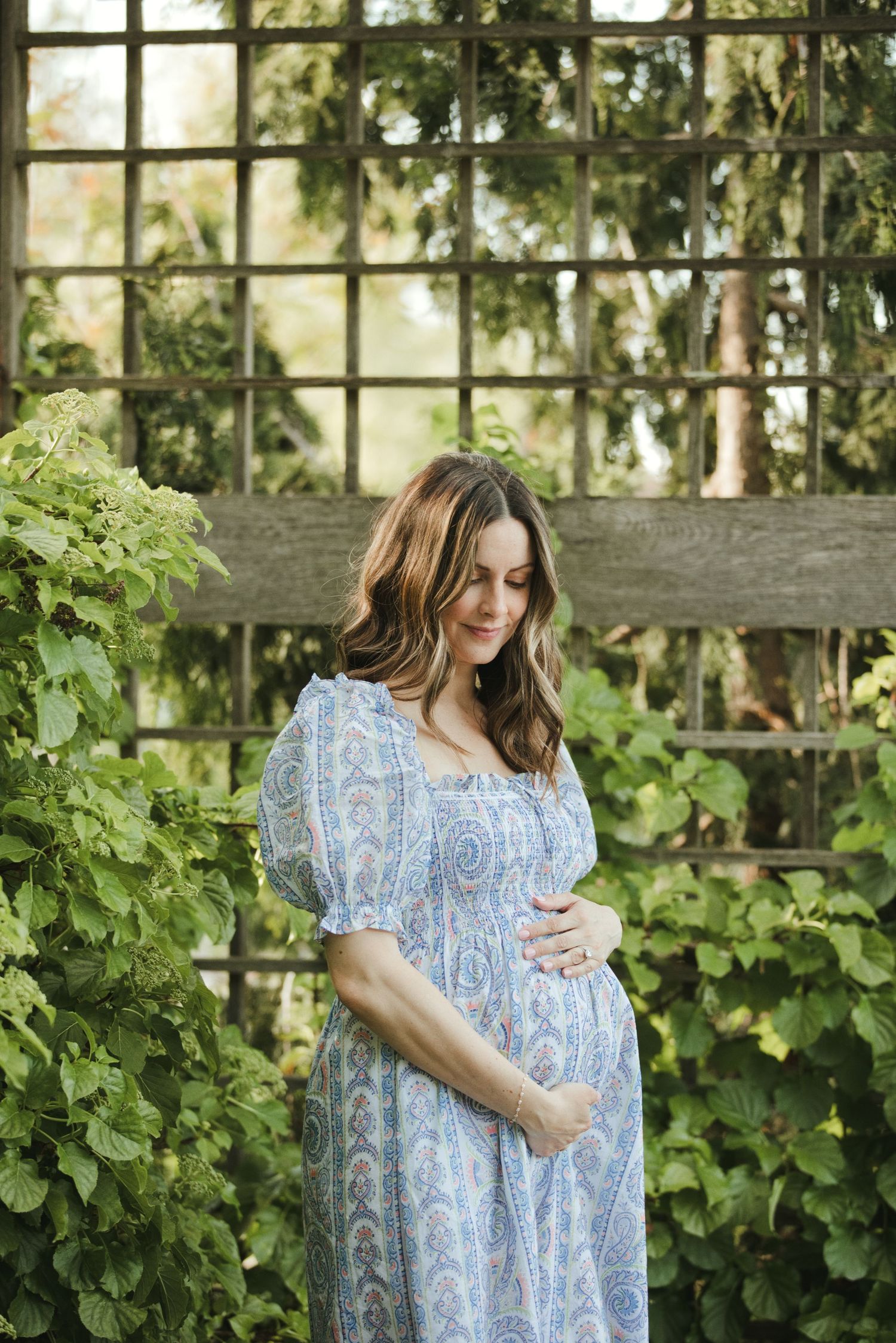 Pregnant woman in blue paisley dress cradling baby bump in lush garden with green trellis backdrop.