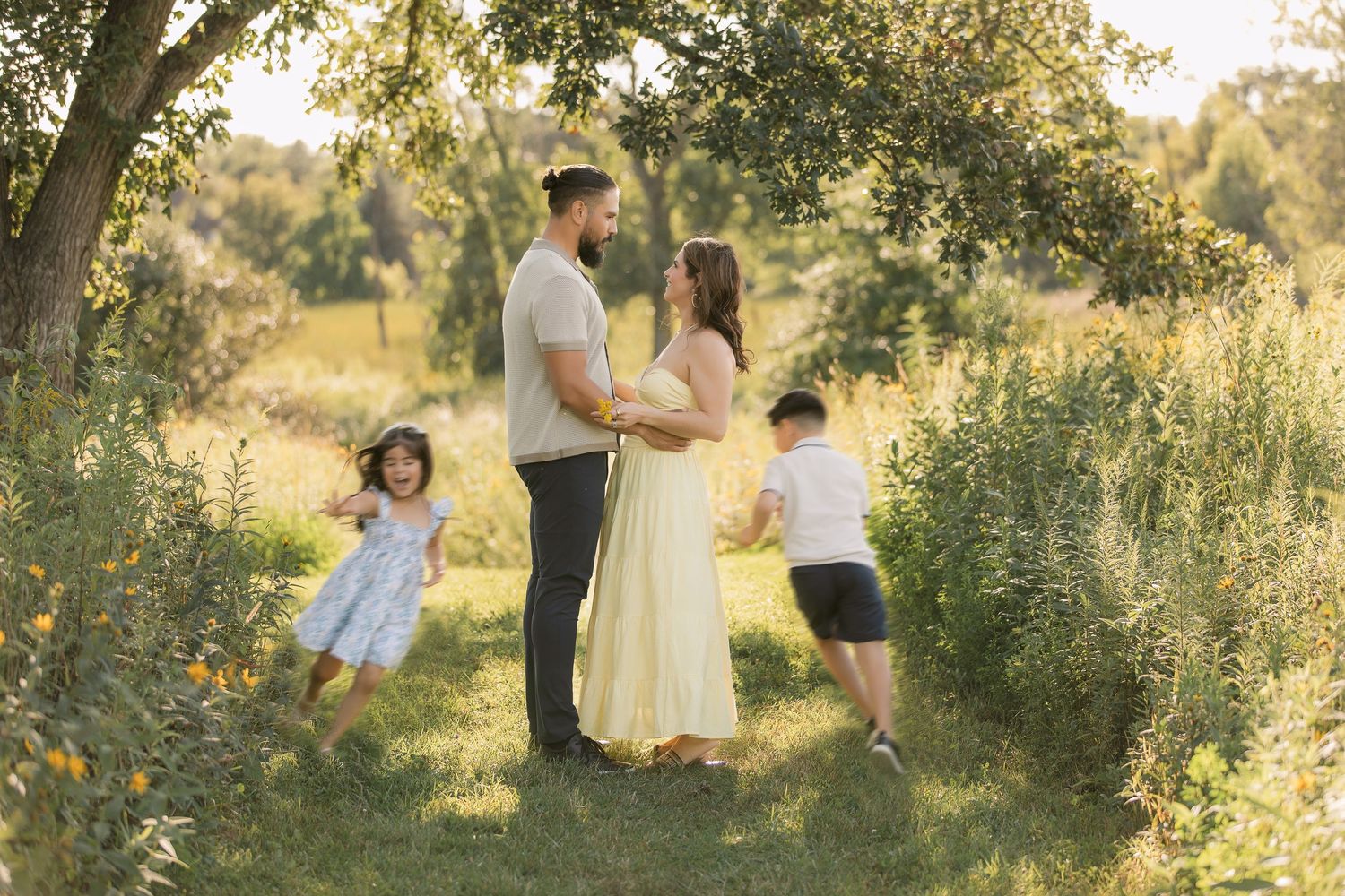 Family of four poses in a sunlit meadow under a tree, parents embracing while two young children play around them.