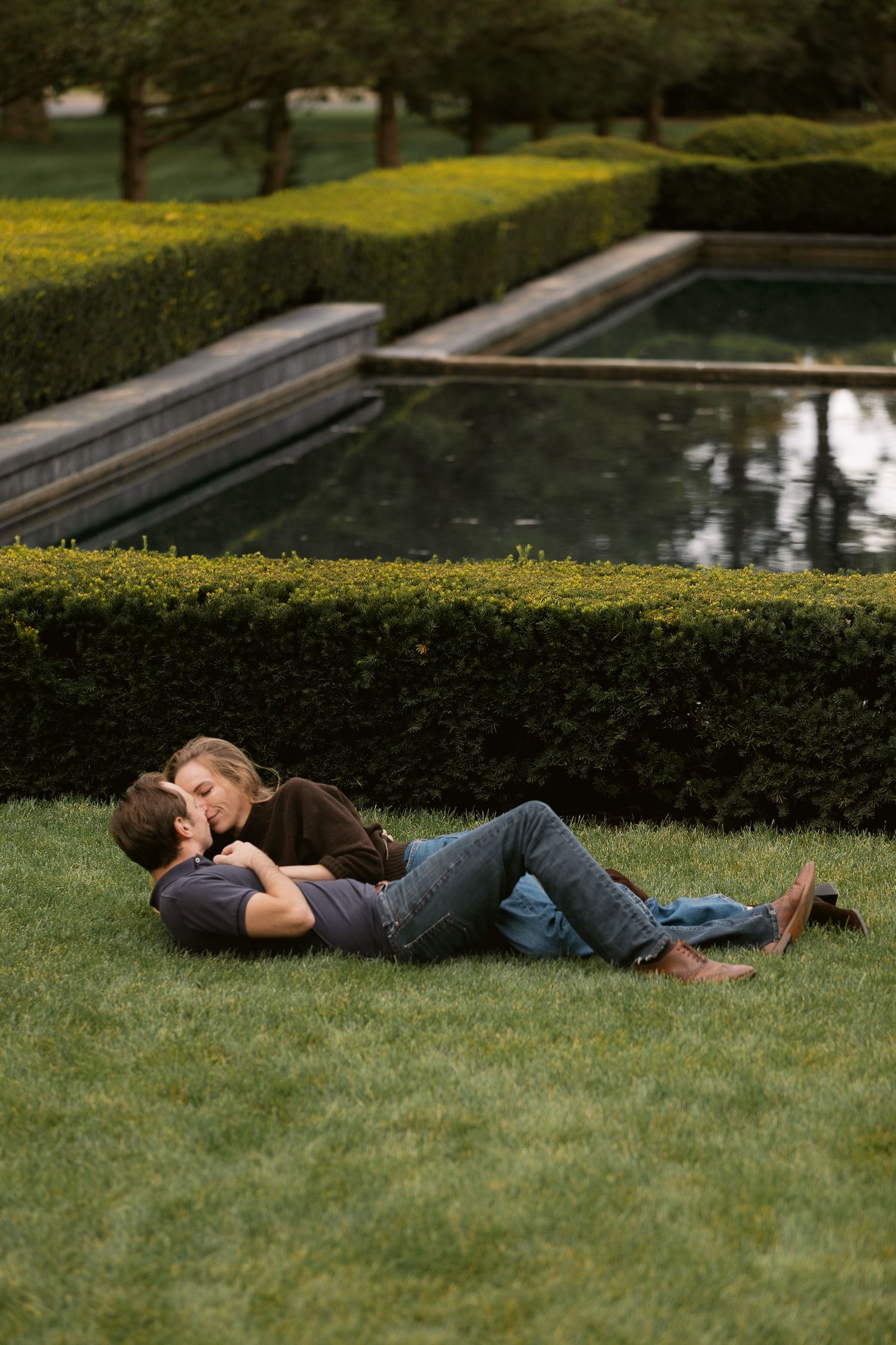 Romantic couple relaxing on lush green grass beside a formal garden pond surrounded by trimmed hedges.