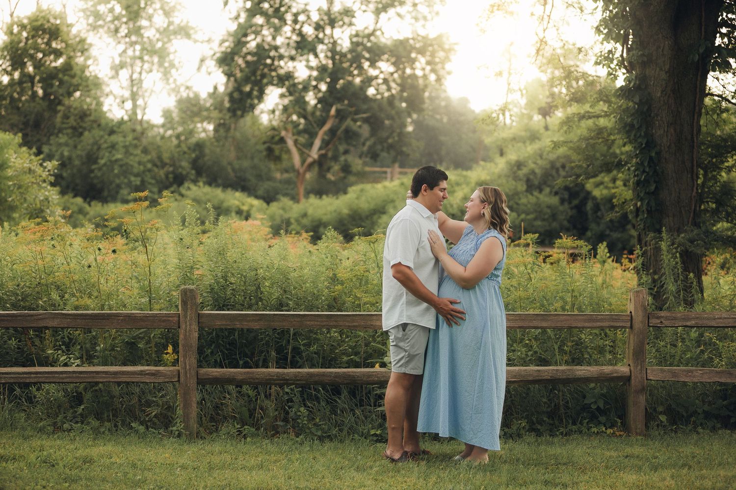 Couple in casual attire shares tender moments during golden hour photoshoot along rustic wooden fence in nature setting.