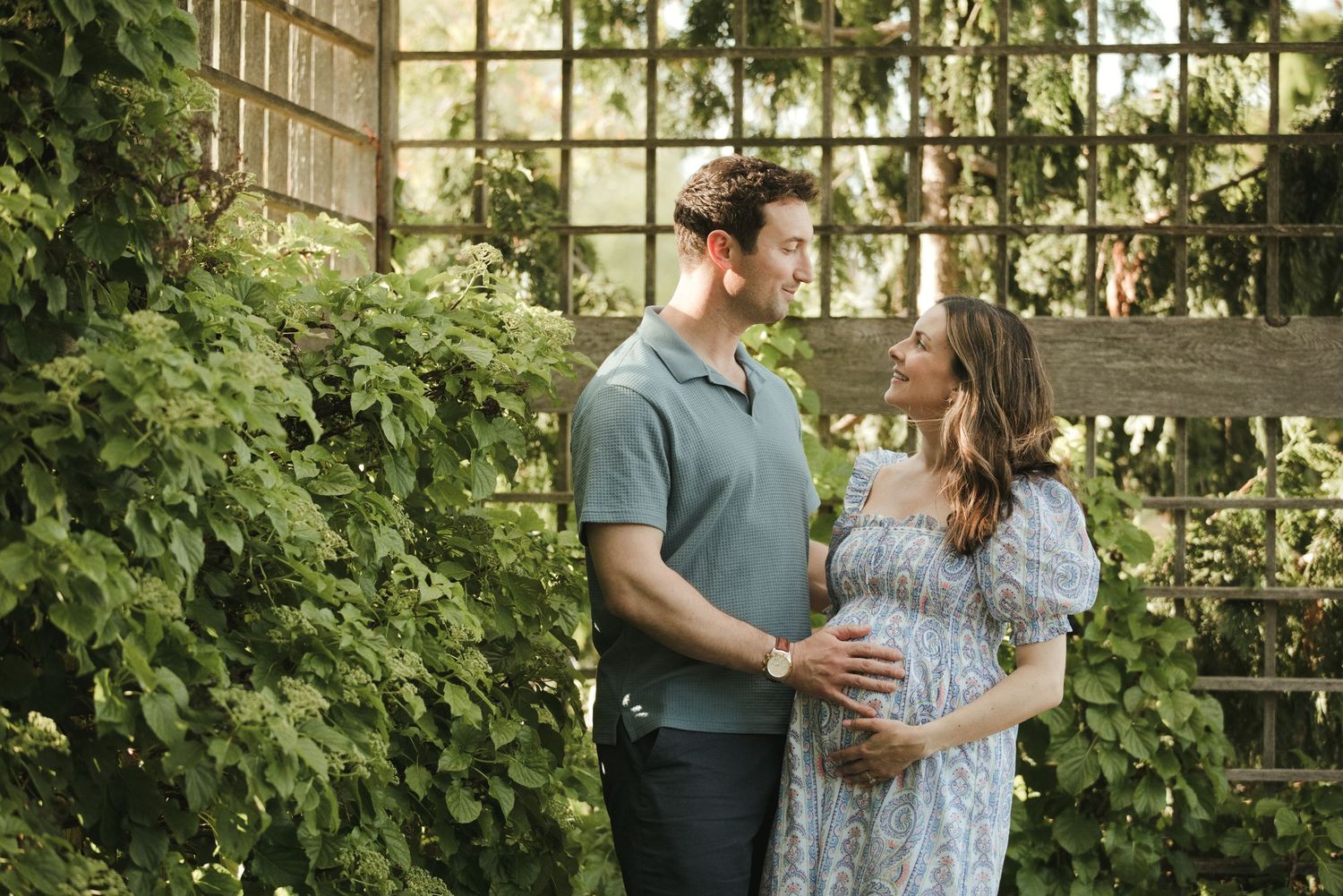 Couple sharing a tender moment in a lush garden, man holding pregnant woman's belly in a floral dress.