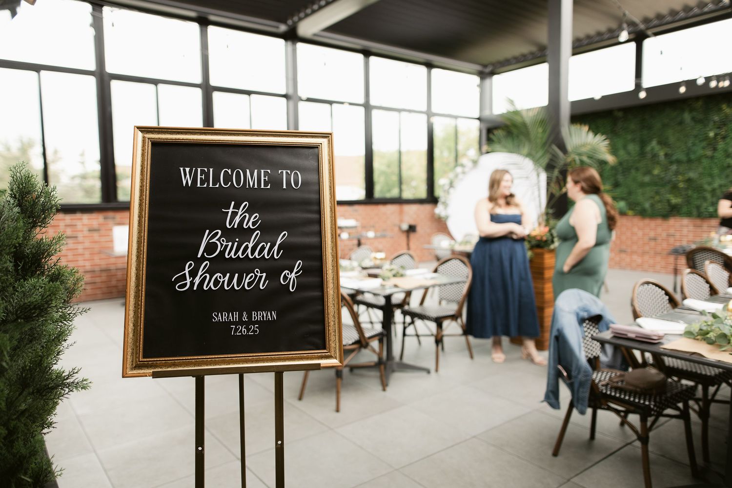 Elegant bridal shower welcome sign in gold frame at an indoor venue with guests mingling in the background.