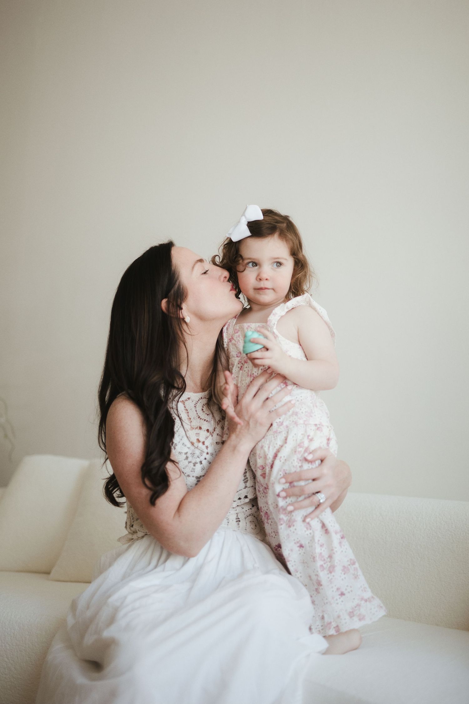 A mother kisses her toddler daughter while sitting on a white couch indoors.