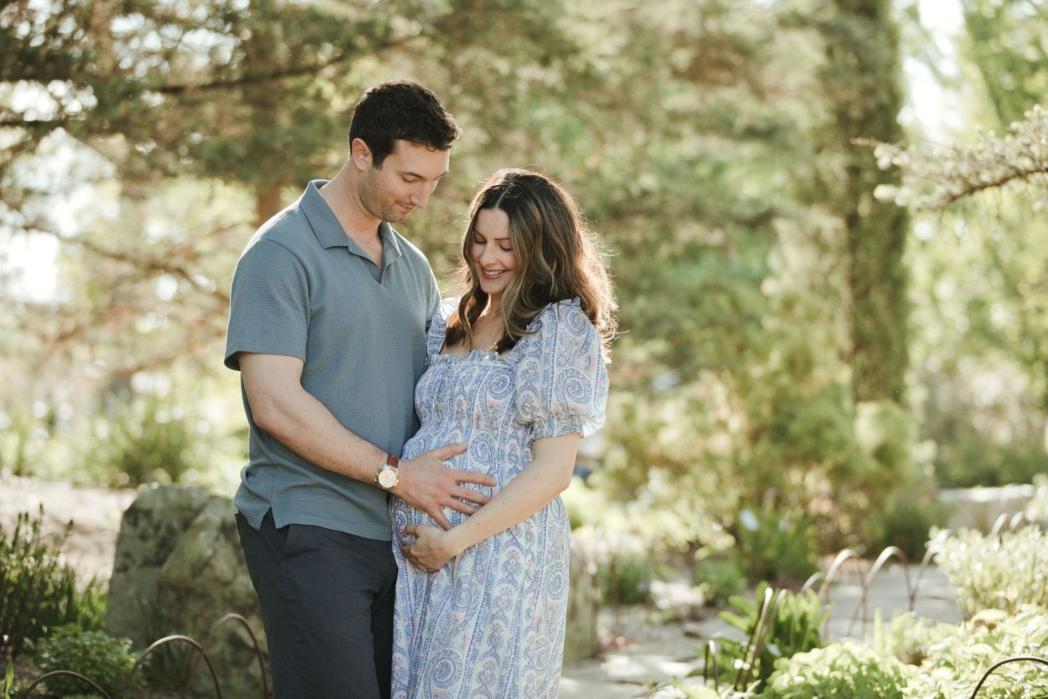Couple smiling at pregnant woman's baby bump outdoors in a sunlit garden during maternity photo session.