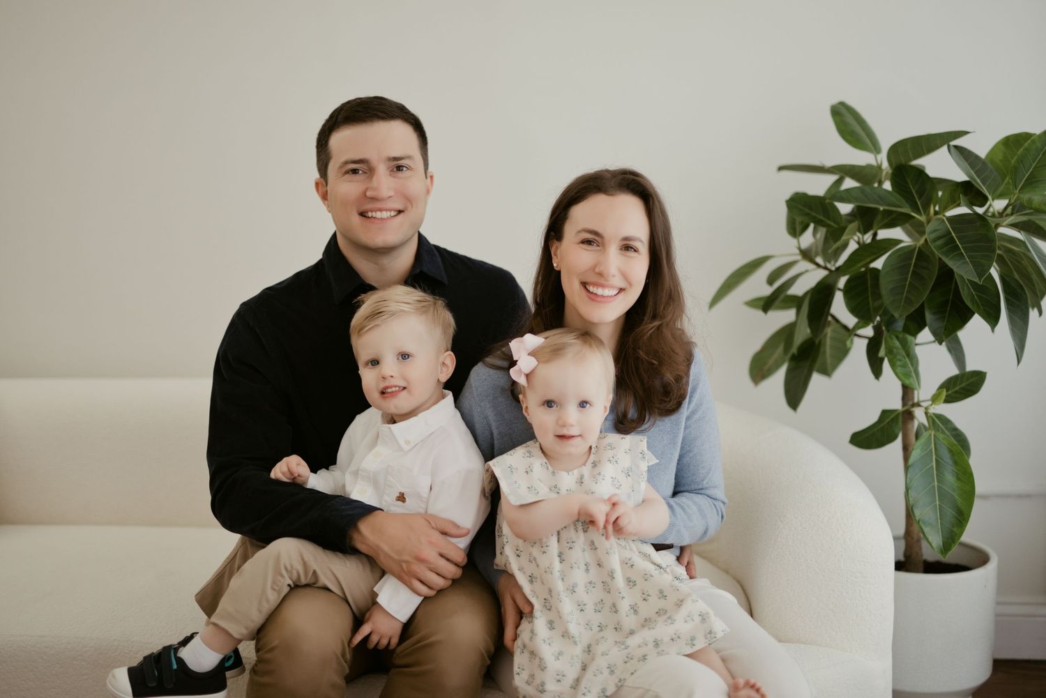 Happy family of four posing on a white sofa, parents smiling with two young toddlers in a bright modern room.