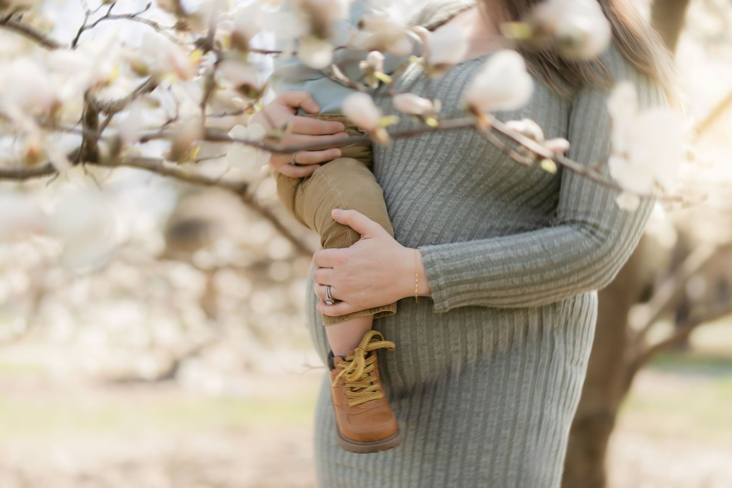 Someone wearing a cozy gray ribbed sweater holds a pair of brown leather boots against a blurred floral background.