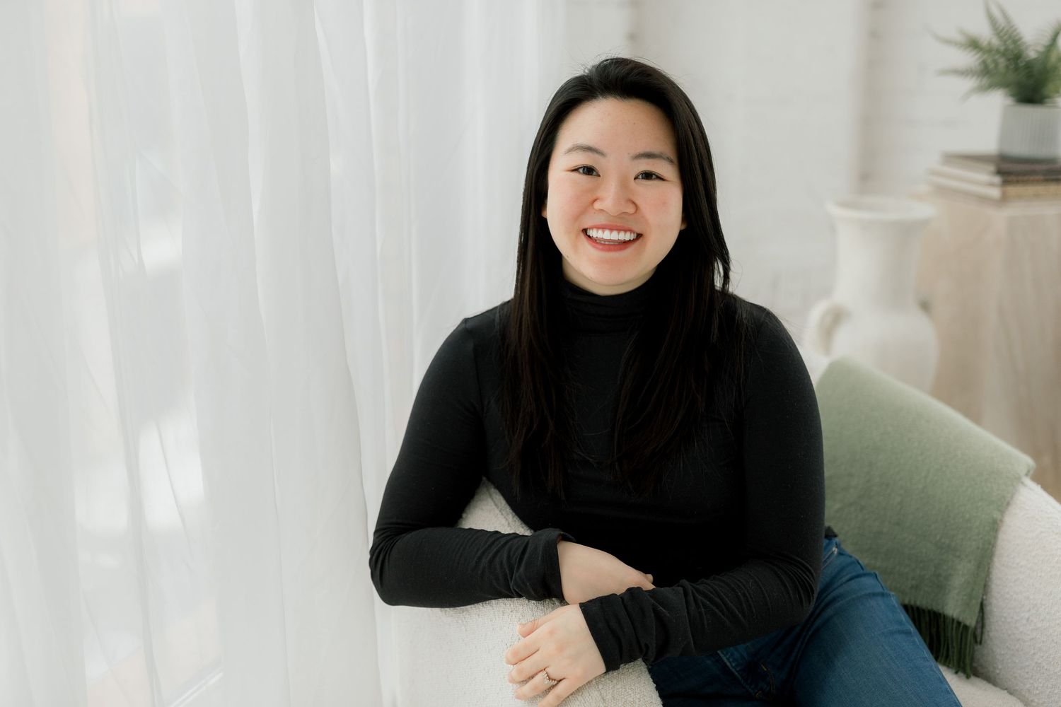 Person in dark outfit sits casually against white backdrop with natural lighting.