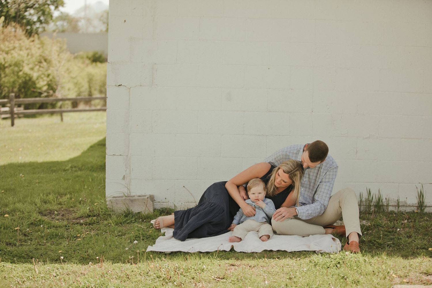 A family of three relaxes on a blanket against a white wall outdoors, with parents cuddling their baby on a sunny day.