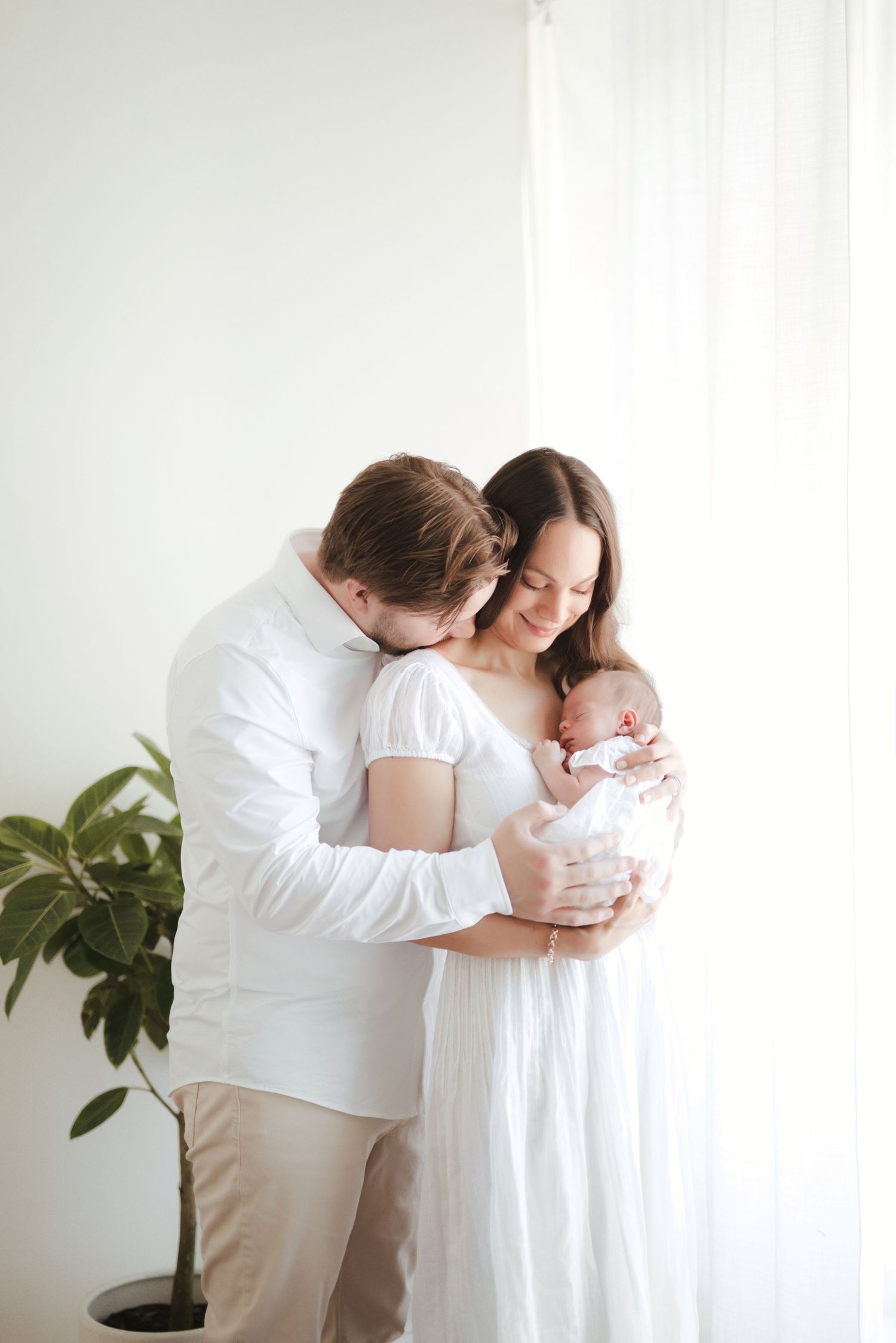 A couple in white clothing tenderly holds their newborn baby near a bright window with a plant nearby.