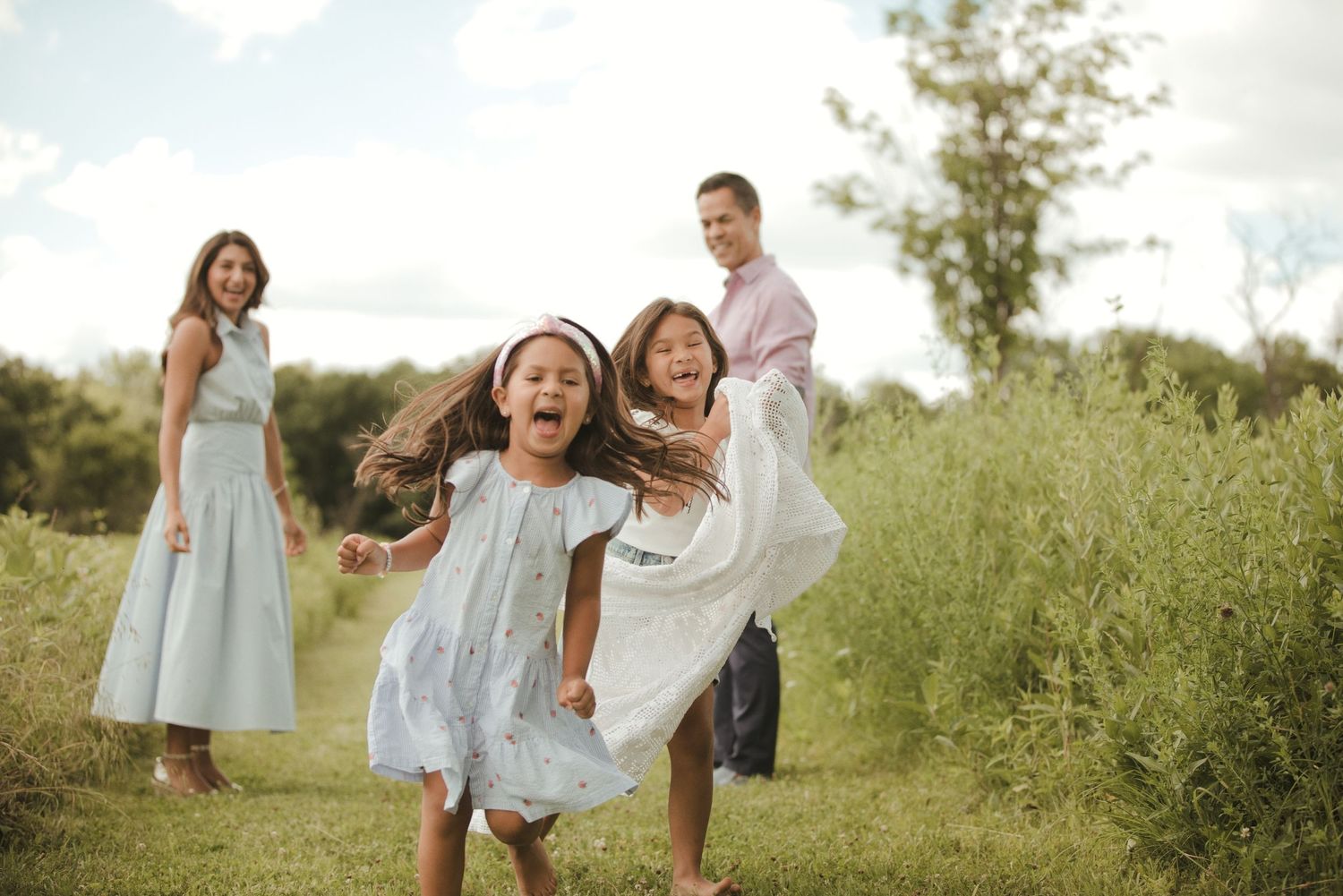 Happy family with two young girls running and laughing outdoors in a green field on a cloudy day.