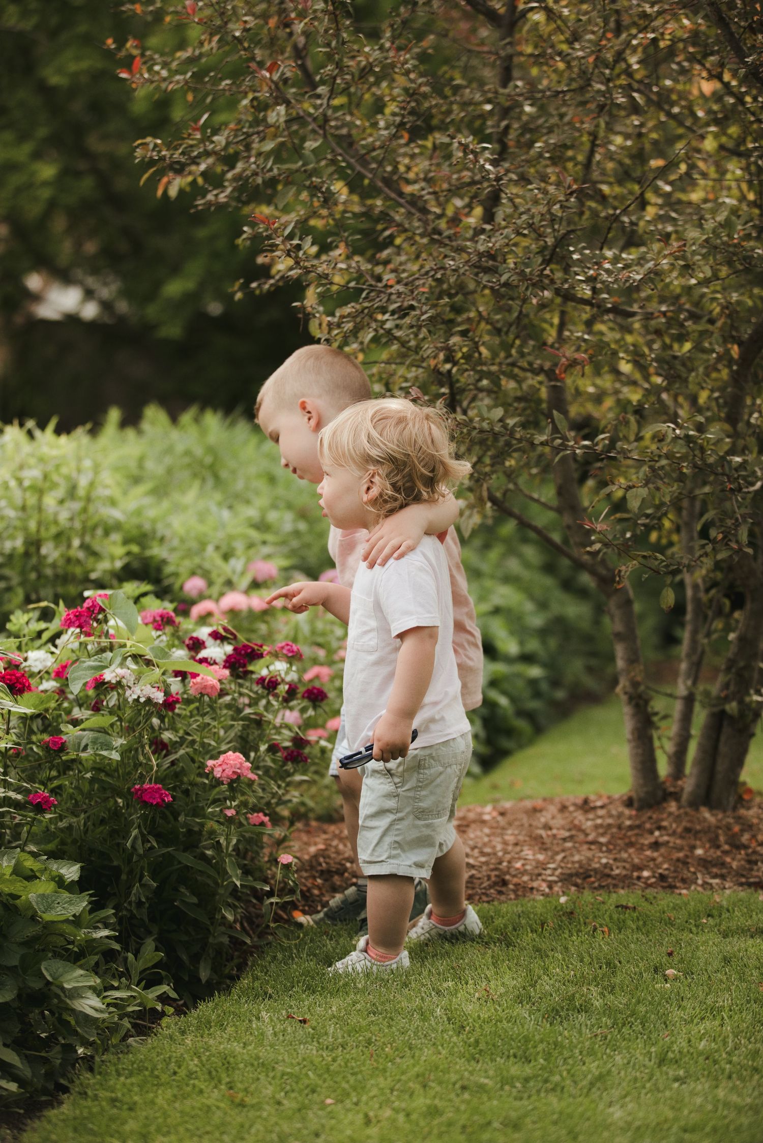 Two toddlers explore a colorful garden flower bed together, with one child hugging the other near blooming pink flowers.