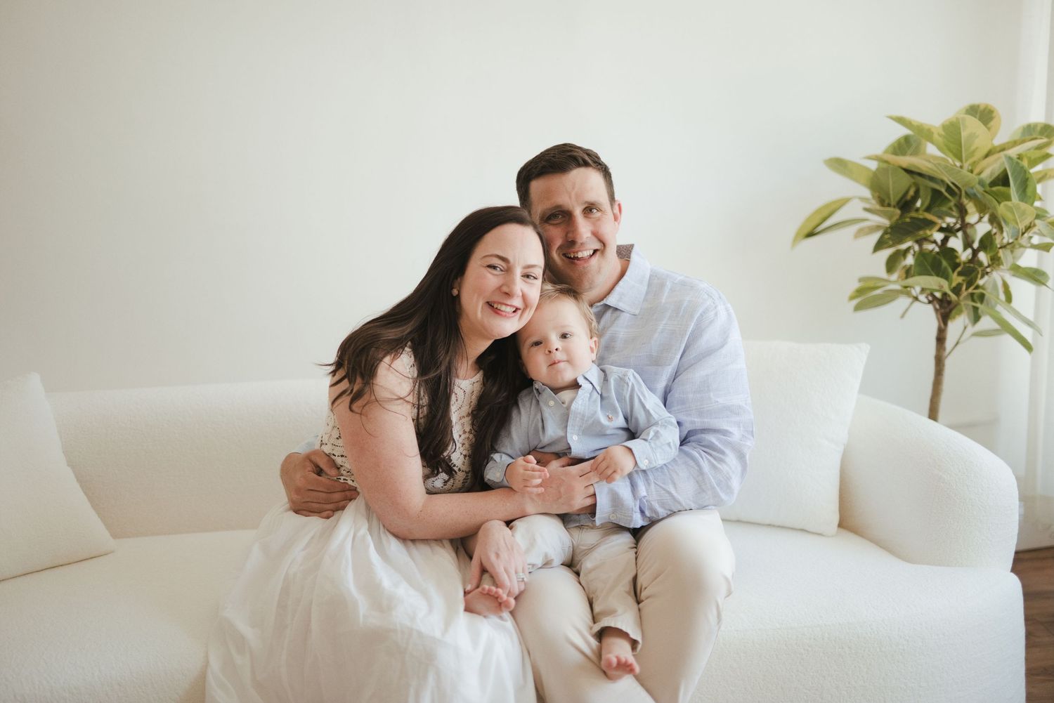 Happy family of three posing on white sofa, parents smiling with baby boy in bright modern living room.